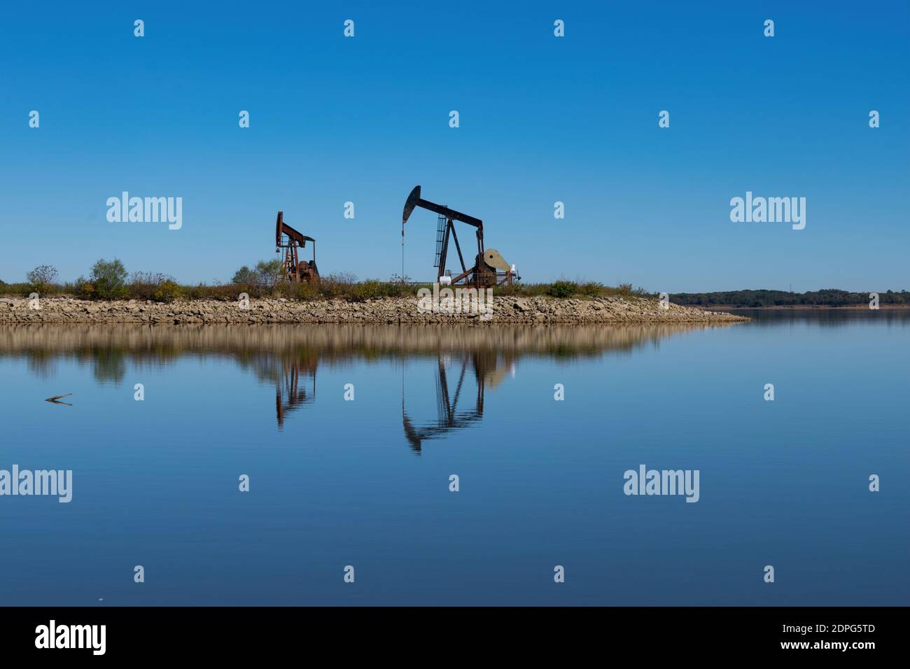 Zwei Ölfeld-Pumpenhebeböcke auf einer felsigen Halbinsel, die mit ihren Reflexen auf der ruhigen, glasigen Wasseroberfläche den See ausragt. Stockfoto