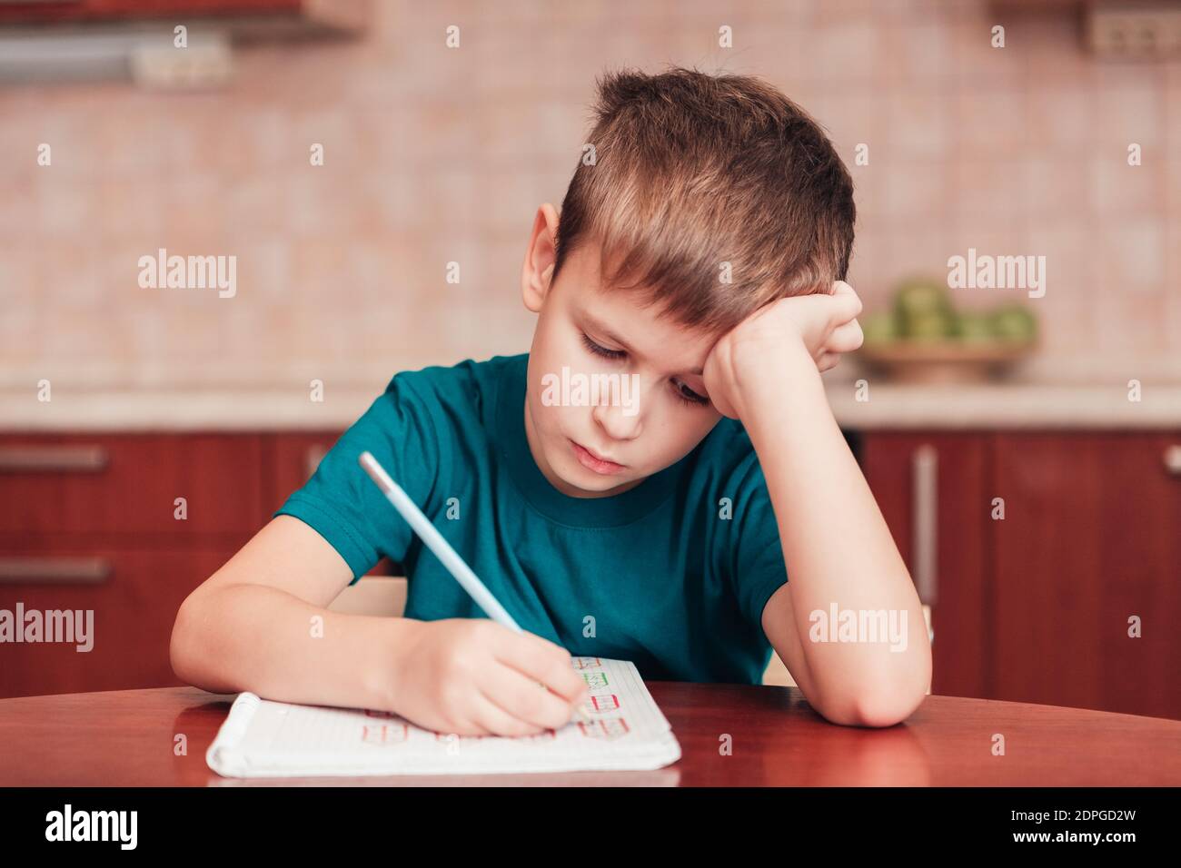 Junge Schuljunge lernen schwer bei Hausaufgaben sitzen von Tisch in Küche mit dem Kopf auf der Hand und Schreibunterricht Im Notebook Stockfoto