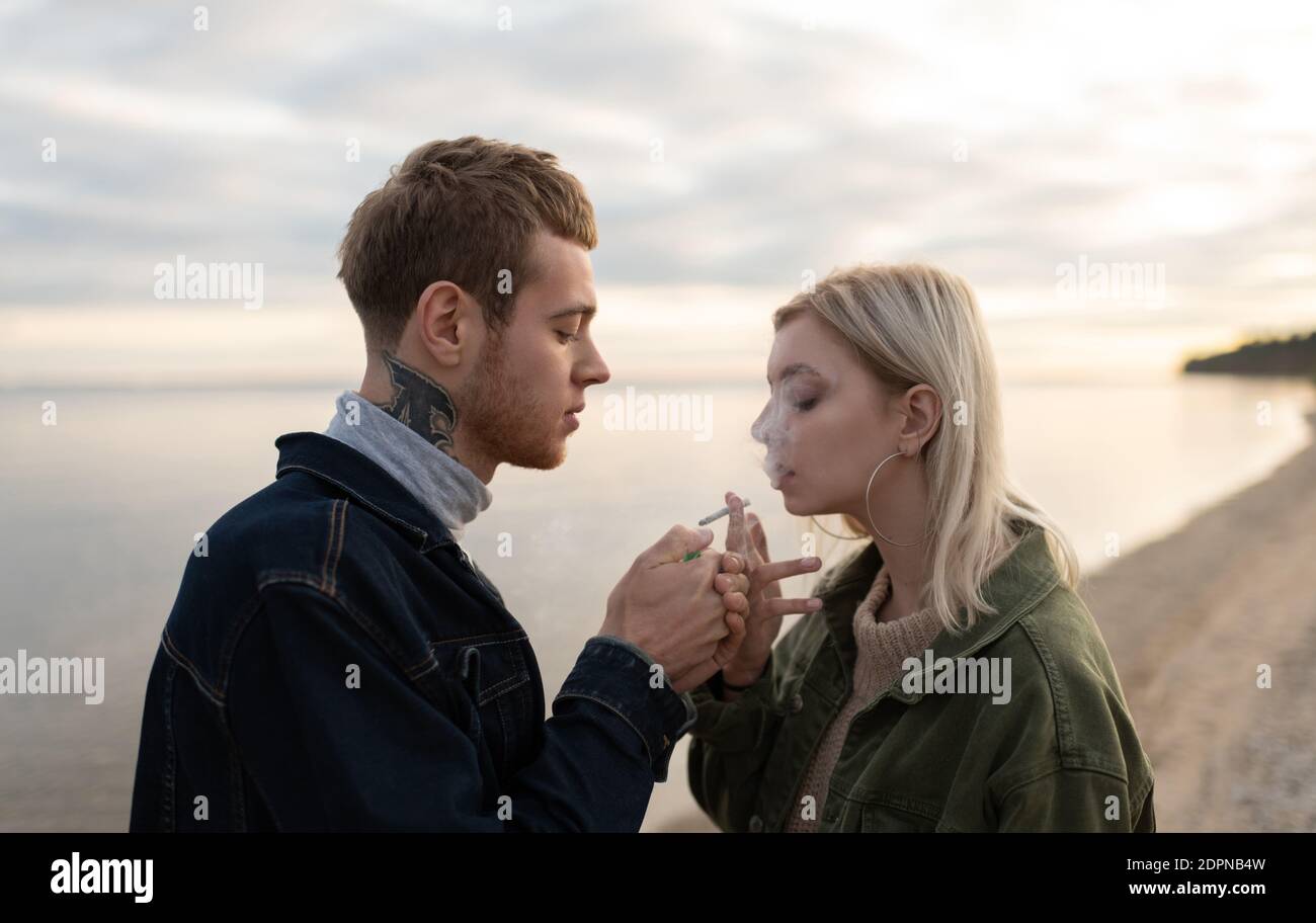 Seitenansicht von jungen Hipstern, die im Stehen Gras rauchen Gegen ruhigen See und bewölkten Himmel während des Datums im Herbst Auf dem Land Stockfoto