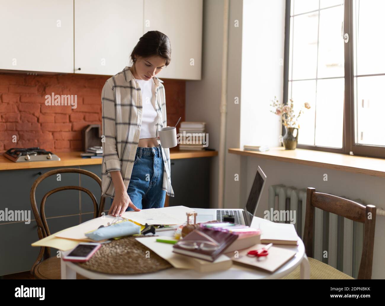 Junge Frau in Hauskleidung mit Kaffee und stehen an Tabelle in der Küche Lesen von Notizen während der Vorbereitung auf die Prüfung Stockfoto