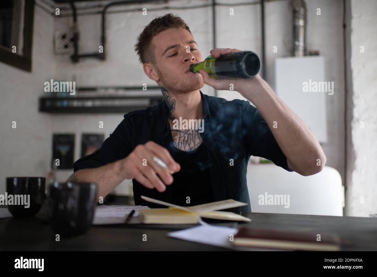 Nachdenklicher junger tätowierter Mann, der Bier aus der Flasche trinkt und raucht Zigarette beim Schreiben am Notebook und beim Sitzen am Tisch Urban Loft Stockfoto