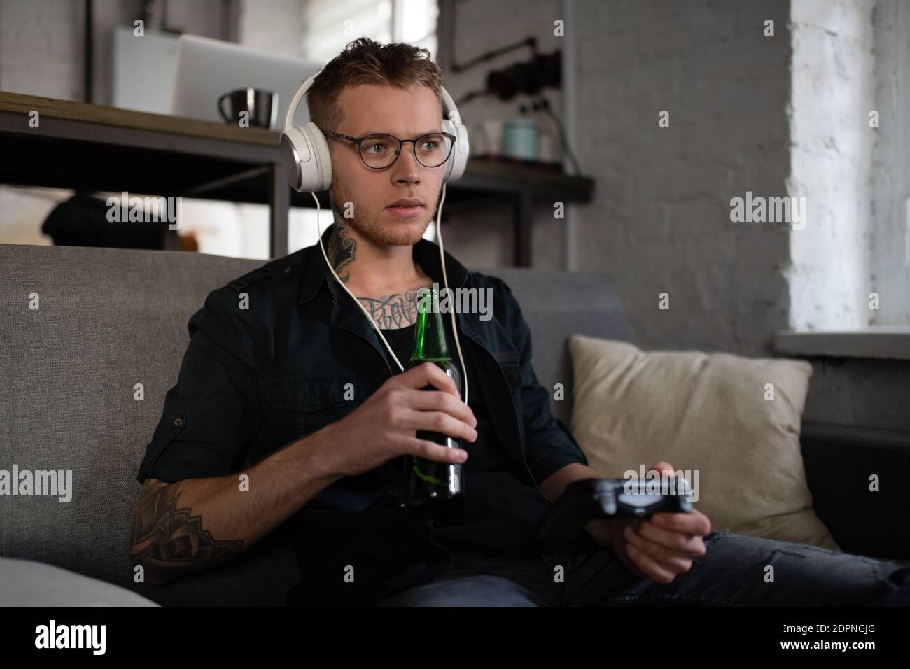 Junger Mann mit Kopfhörer und Gläsern, der Bier aus der Flasche trinkt Und mit Controller, um Videospiel spielen, während entspannen auf Sofa zu Hause Stockfoto