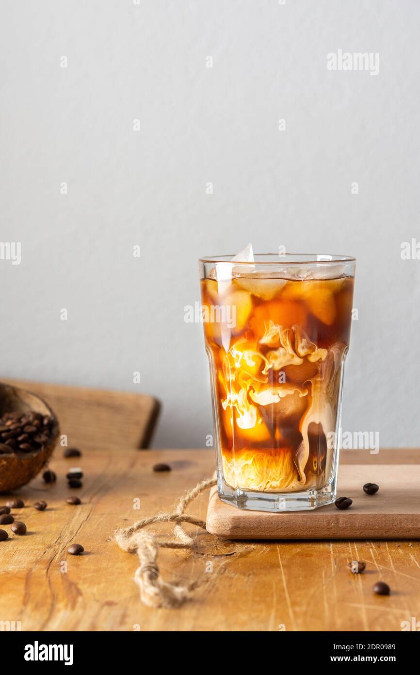 Kalter erfrischender Kaffee mit Eis und cremigen Wolken in einem Glas auf einem Holzbrett Nahaufnahme, Kakaobohnen in Kokosnussschale Stockfoto