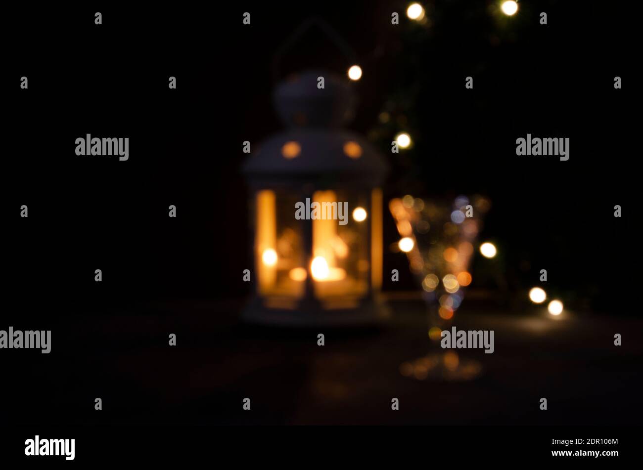 Glänzend verschwommener Hintergrund für Design. Glas mit Weißwein mit Girlande Lichter und Kerze Laterne verschwommen. Nachtbild. Wunderschöne Beleuchtung. Stockfoto