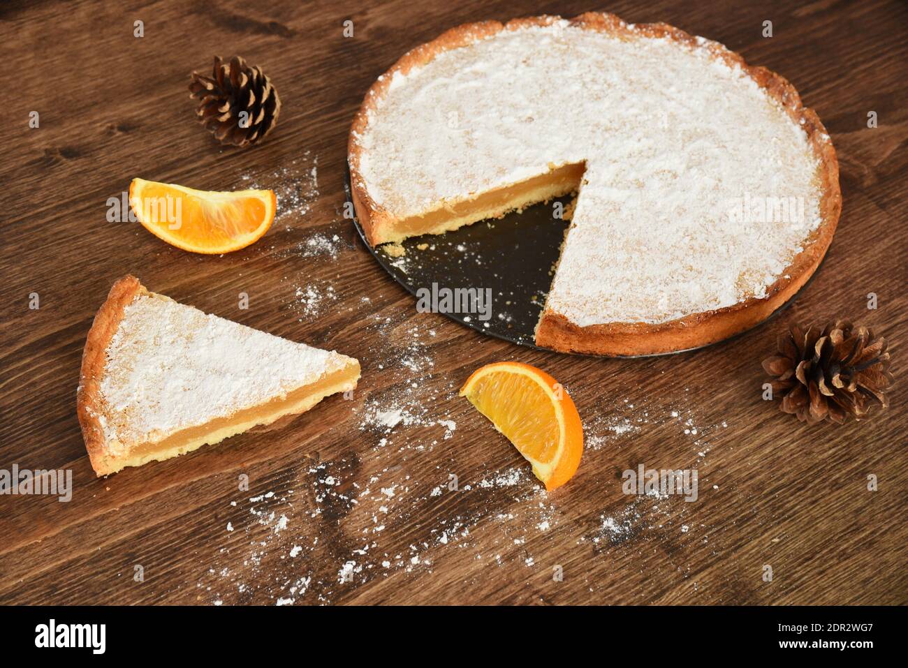 Stück hausgemachter Kuchen mit Zitronen- und Orangenfüllung, bedeckt mit Puderzucker auf einem Holztisch in der Nähe von Orangenfrüchten und Tannenzapfen Stockfoto