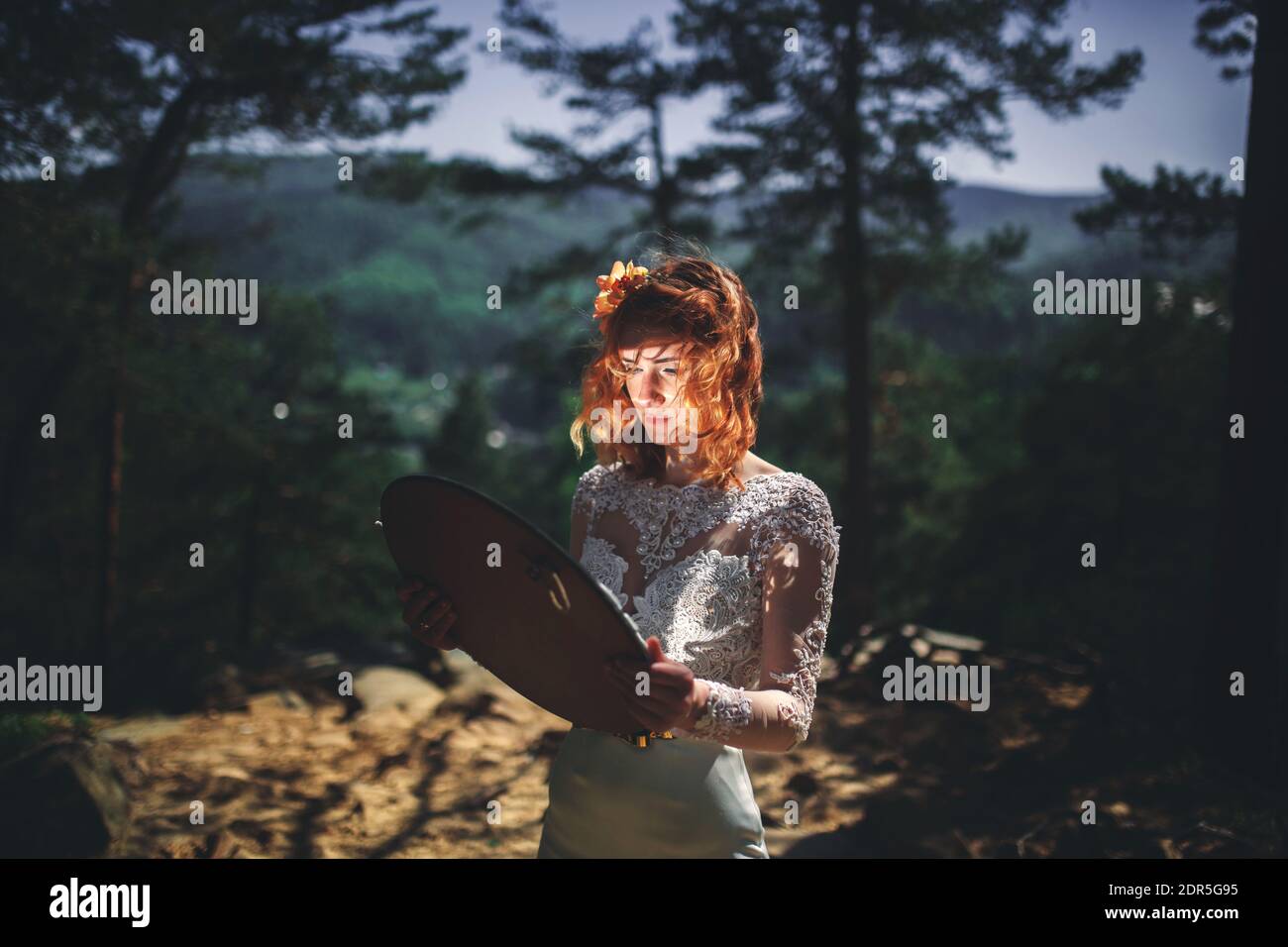 Schöne junge rothaarige Braut im Wald mit einem Blumenkranz auf dem Kopf und Spiegel. Frau in langen weißen Kleid im Freien am Sommertag. Hochzeit Stockfoto