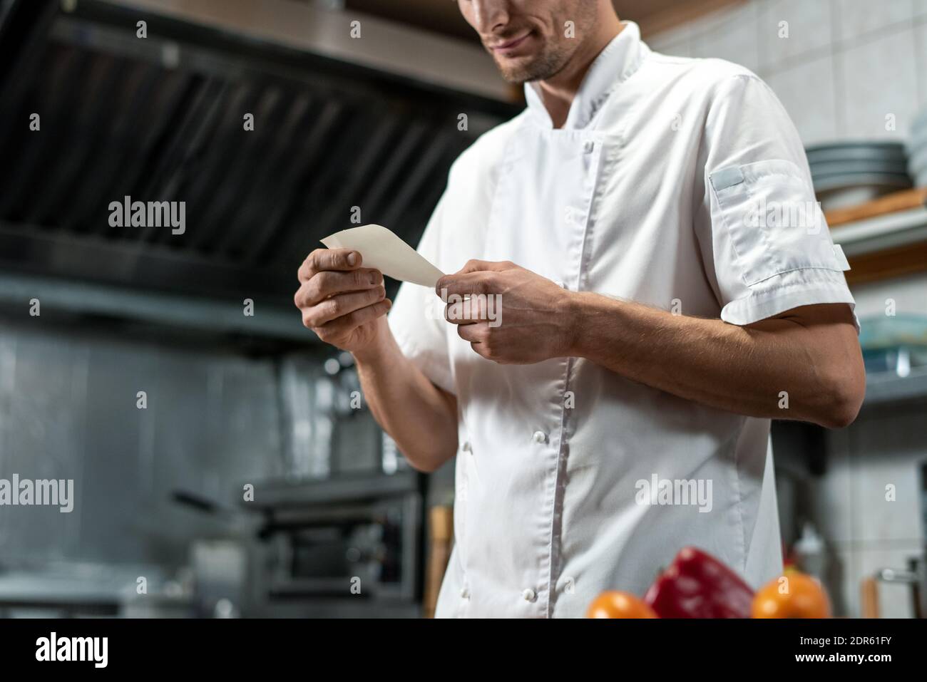Junger Koch in weißer Uniform schaut sich die Liste der Zutaten an Für Gemüsesalat oder andere Gerichte beim Kochen Die Küche Stockfoto