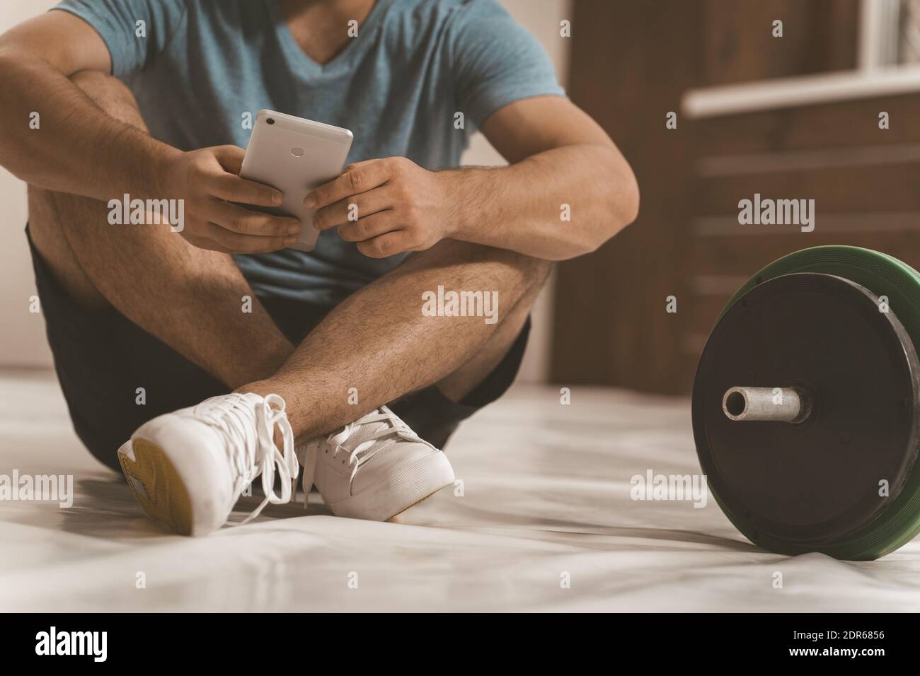 Keine Gesichtsaufnahme eines Fitnesstrainers, der online sucht oder auf dem Boden mit Smartphone sitzt, schwarze und grüne Fitness-Langhantel, Geräte Stockfoto