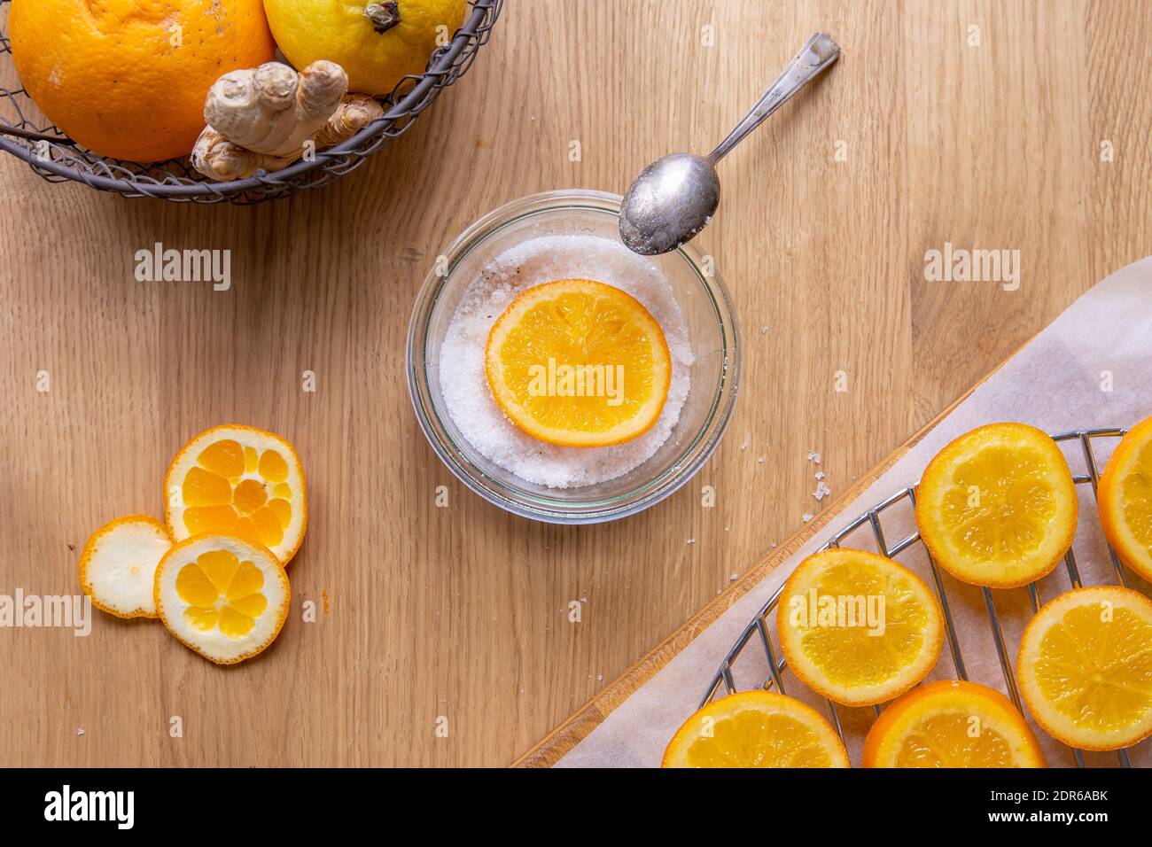 Kandierte Orangenscheiben herstellen: Holztisch mit trocknenden Orangenscheiben, die nach dem Kochen in Sirup getrocknet werden, und eine Schüssel mit Zucker und einer Orangenscheibe. Stockfoto