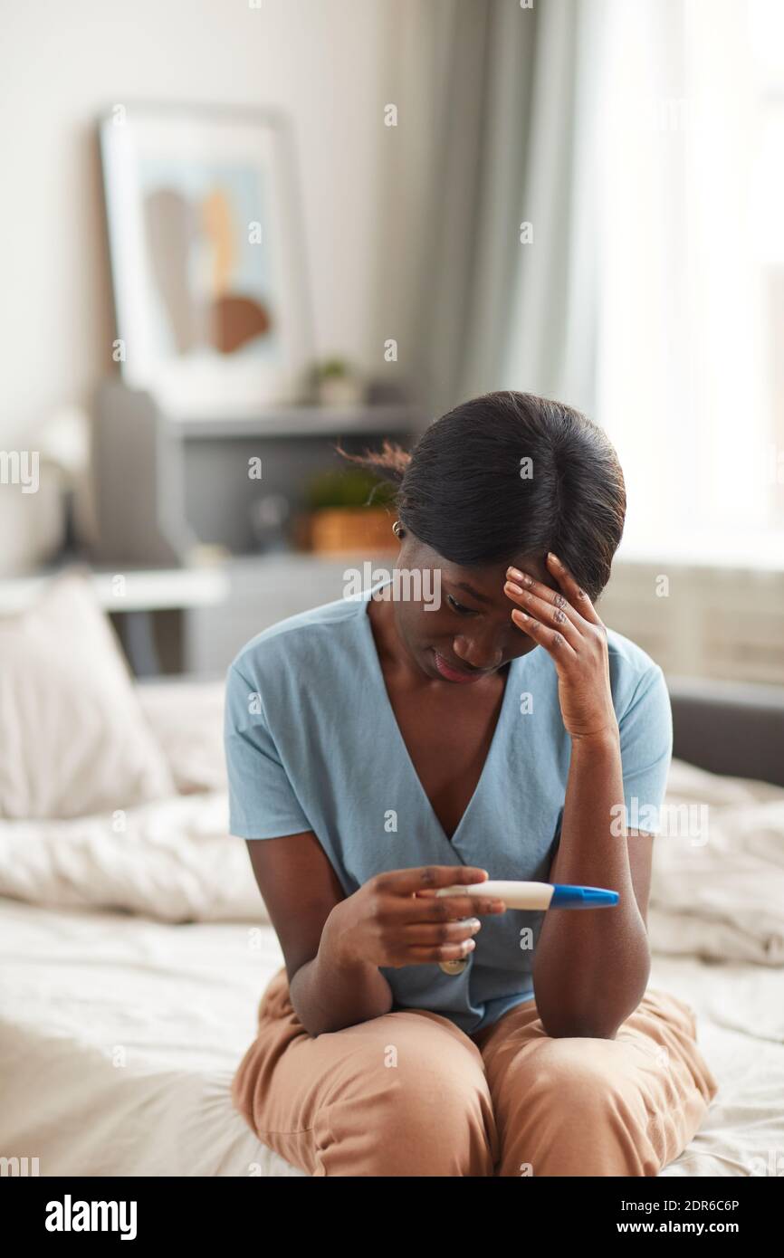 Vertikales Porträt einer besorgten afroamerikanischen Frau, die einen Schwangerschaftstest ansieht Während Sie zu Hause auf dem Bett sitzen Stockfoto