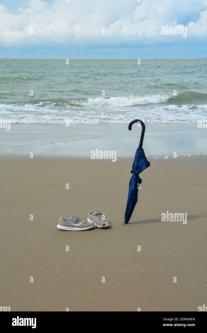 Ein geschlossener Schirm ist im Sand stecken und alt Schuhe mit Löchern  stehen vor dem Strand Das Meer mit vielen Wellen Stockfotografie - Alamy