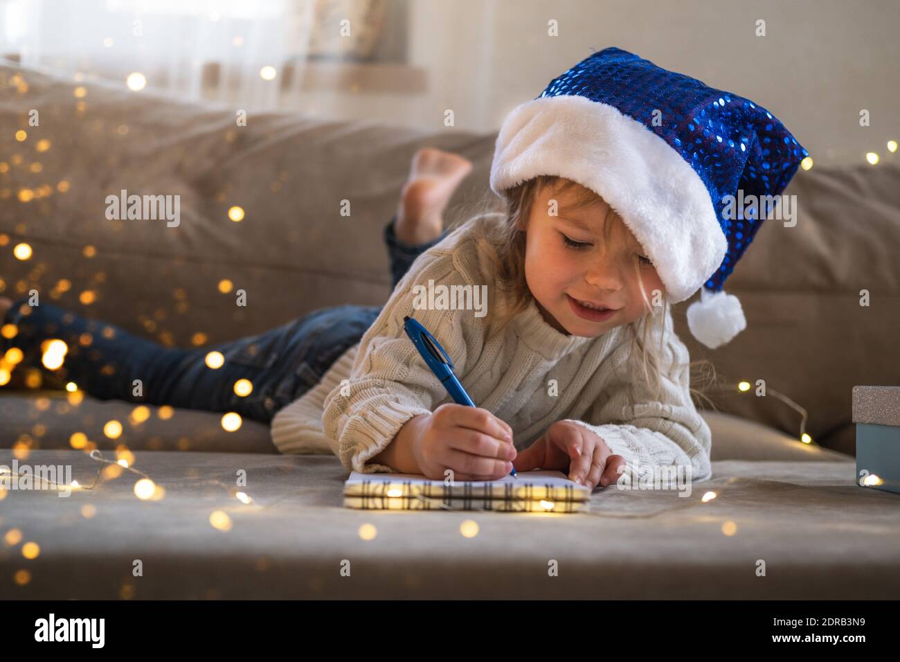 Junges Mädchen in blauem Weihnachtsmütze, das einen Wunschbrief an den Weihnachtsmann schreibt. Weihnachten oder Neujahr Konzept. Stockfoto