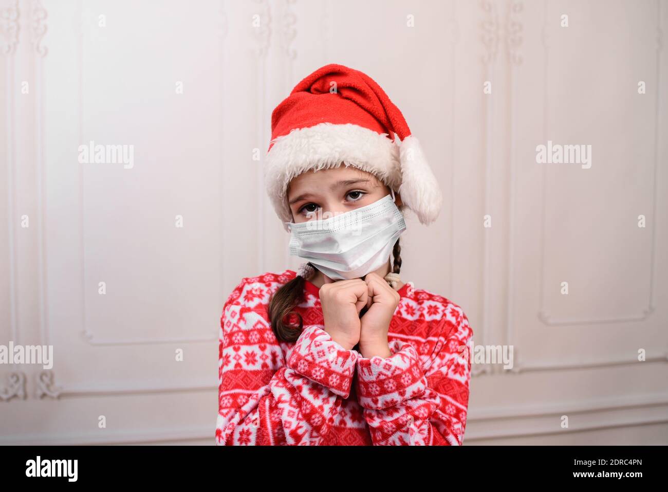 Deprimiertes kleines Mädchen, das traurig aussieht und einen Weihnachtsmann Hut und eine medizinische Maske trägt. Kind Mädchen mit unglücklichem Ausdruck am weihnachtstag. Modellversion. Stockfoto
