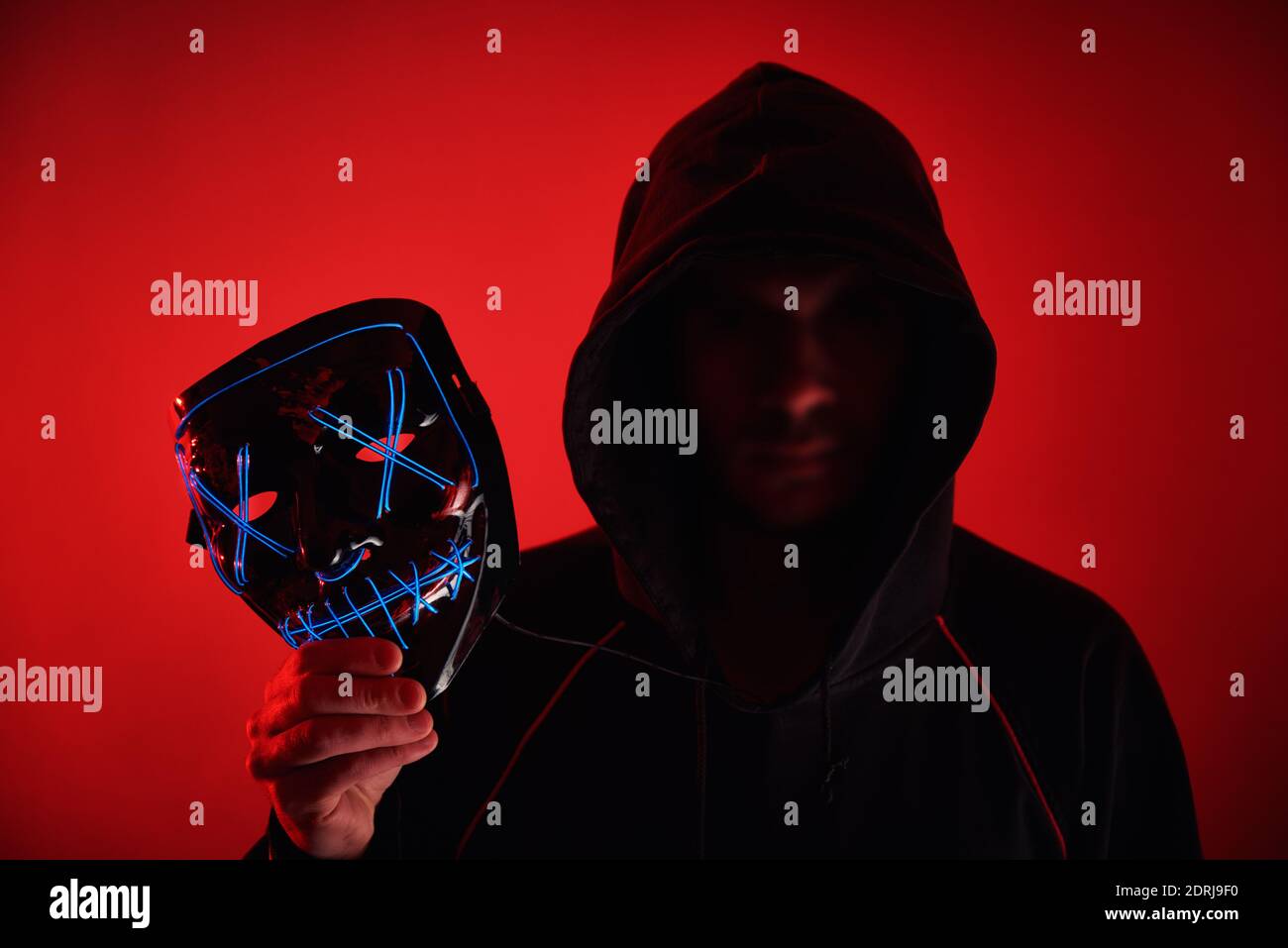 Mann im Hoodie mit verschwommenem Gesicht hält Neon glühende Gruselmaske. Anonymes Konzept Stockfoto