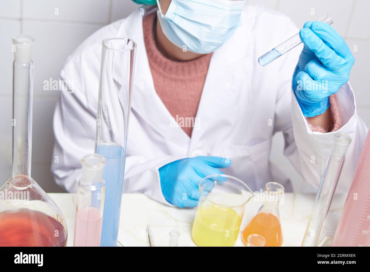 Labortechniker in weißer Schicht, blauen Handschuhen, Schutzkappe und Gesichtsmaske mit einem Reagenzglas, das Tests mit verschiedenen Reagenzgläsern durchführt Stockfoto