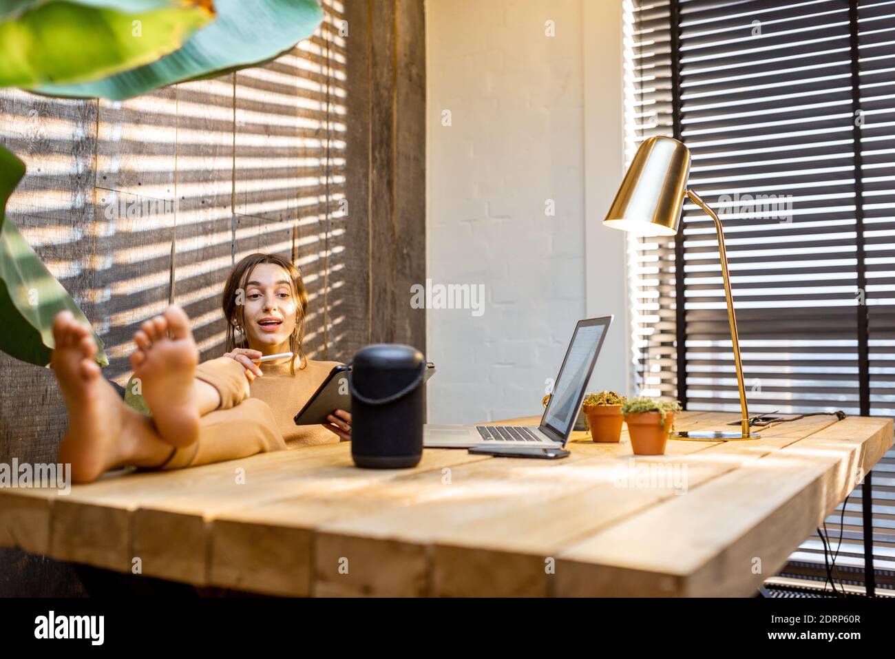 Frau, die mit dem intelligenten Lautsprecher spricht, während sie am digitalen Tablet im gemütlichen Heimbüro arbeitet. Konzept eines Smart Home und Künstliche Intelligenz Stockfoto