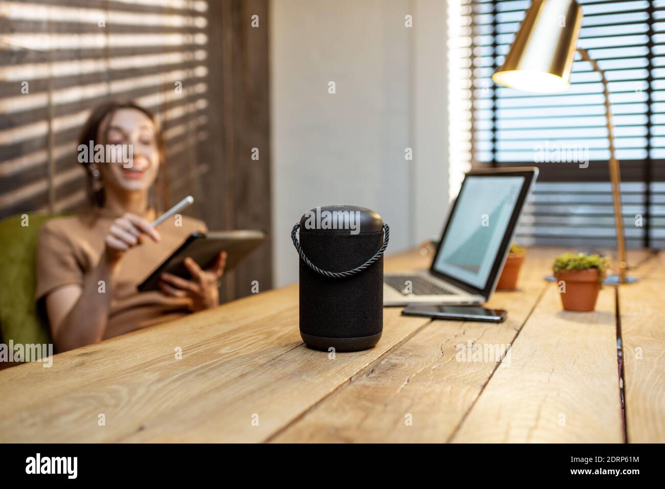Frau, die mit dem intelligenten Lautsprecher spricht, während sie am digitalen Tablet im gemütlichen Heimbüro arbeitet. Konzept eines Smart Home und Künstliche Intelligenz Stockfoto