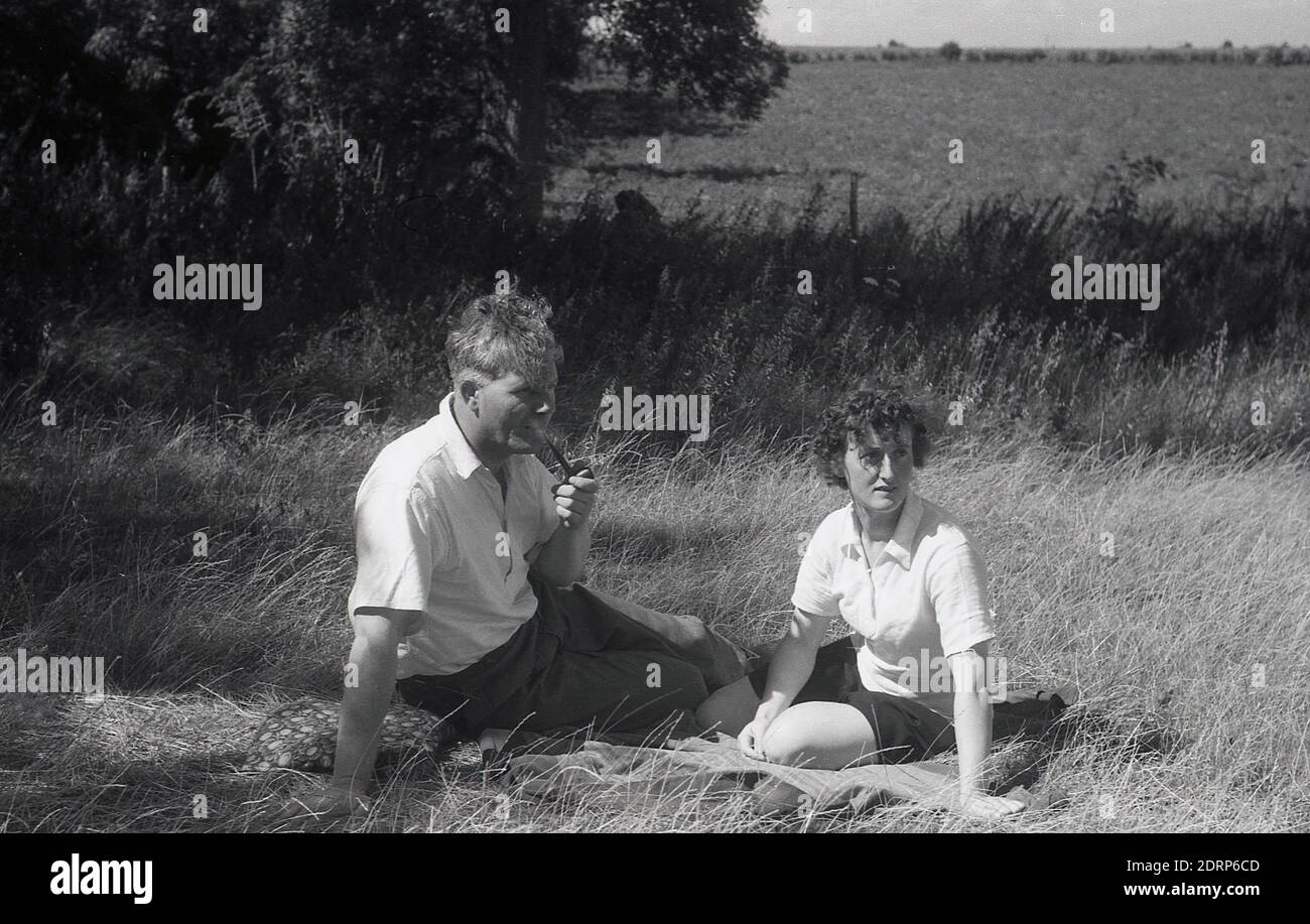 1950er Jahre, historisch, Sommer im ländlichen Kent, ein Paar, das auf einem Teppich auf einem langen Grasfeld auf dem Land sitzt, der Gentleman raucht eine Pfeife, Kent, England, Großbritannien. Stockfoto