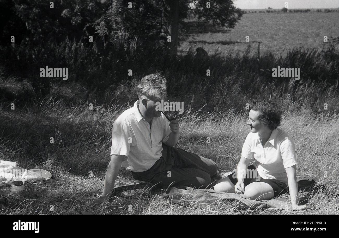 1950er Jahre, historisch, Sommerzeit und ein Picknick im ländlichen Kent, ein Paar sitzt zusammen auf einem Teppich auf einem Feld auf dem Land, der Herr raucht eine Pfeife, Kent, England, Großbritannien. Stockfoto