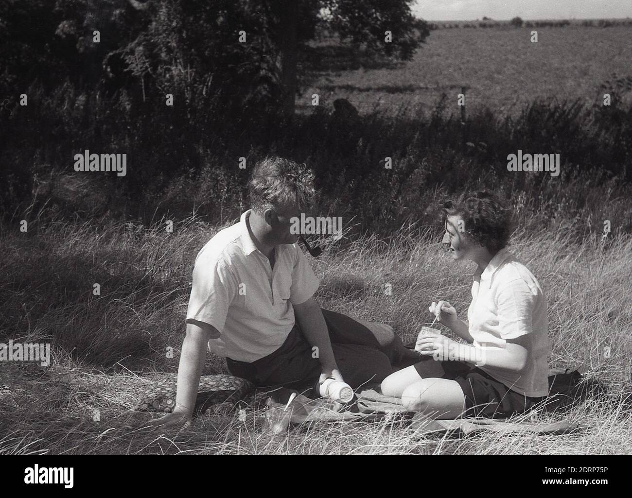 1950er Jahre, historisch, Sommerzeit und ein Picknick im ländlichen Kent, ein Paar sitzt zusammen auf einem Teppich auf einem Feld auf dem Land, der Herr raucht eine Pfeife, Kent, England, Großbritannien. Stockfoto