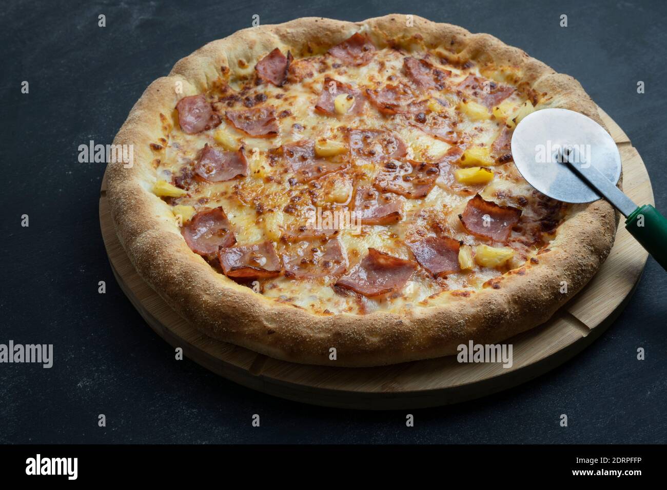 Leckere hawaiianische Pizza (Käse, knuspriger Speck und Ananas) und Pizza Cutter wheel.Dark Hintergrund mit Copy Space. Stockfoto