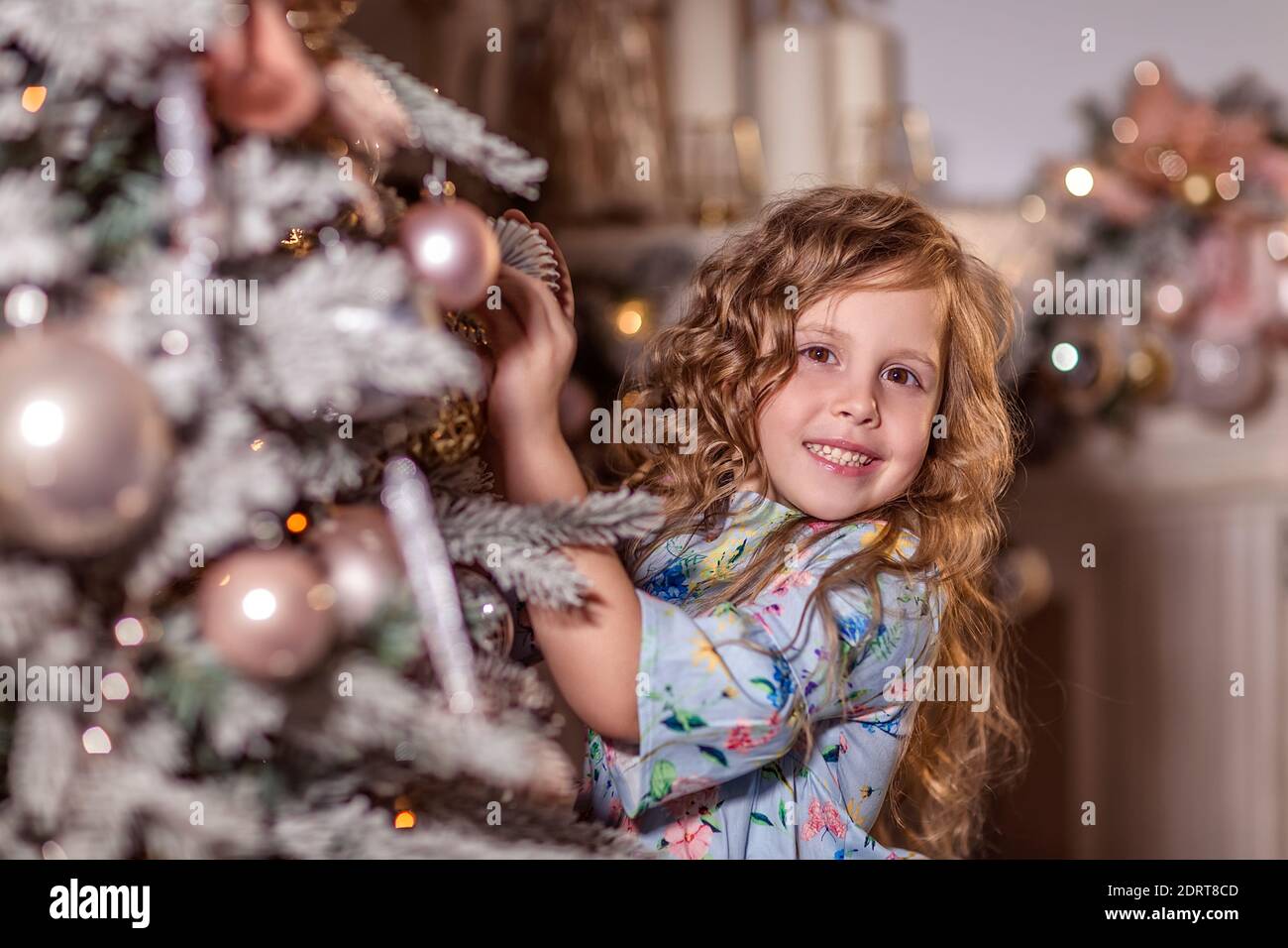 Ein schönes lachendes Mädchen mit lockigen langen Haaren steht neben einem geschmückten Weihnachtsbaum. Schmückt den Weihnachtsbaum. Hängt Luftballons und Spielzeug auf dem CH Stockfoto