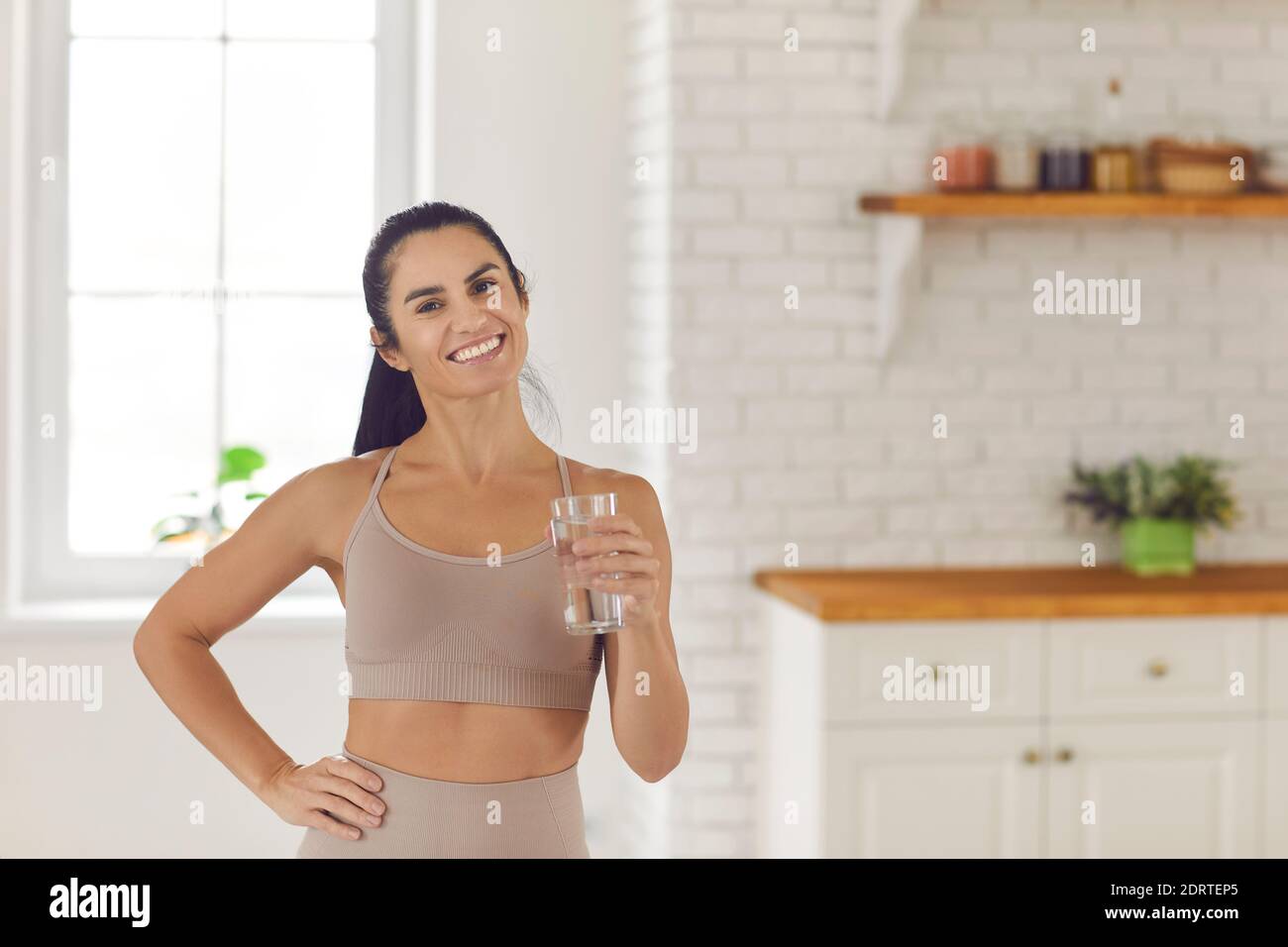 Lifestyle-Porträt einer Fitness-Frau, die ein Glas Wasser hält, während sie in der Küche steht. Stockfoto