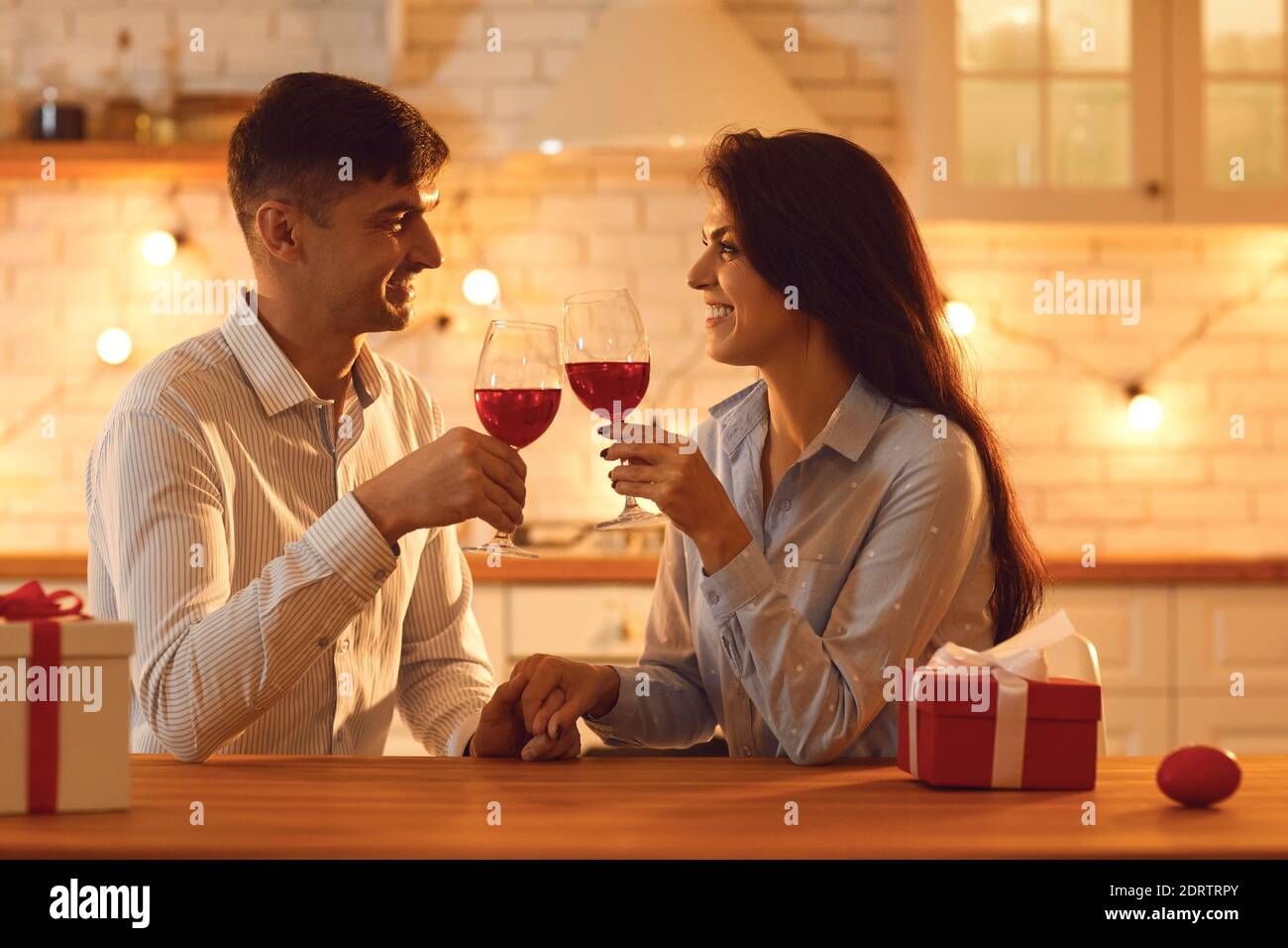 Lächelndes liebevolles Paar, das mit Gläsern Wein sitzt und die Hände während der Feier des Valentinstag hält Stockfoto