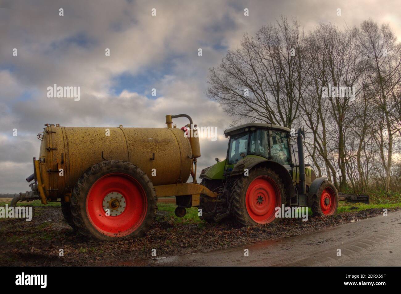 Gelber Gülletanker und grüner Traktor mit roten Rädern in Schlamm unter blauem Himmel mit Wolken Stockfoto