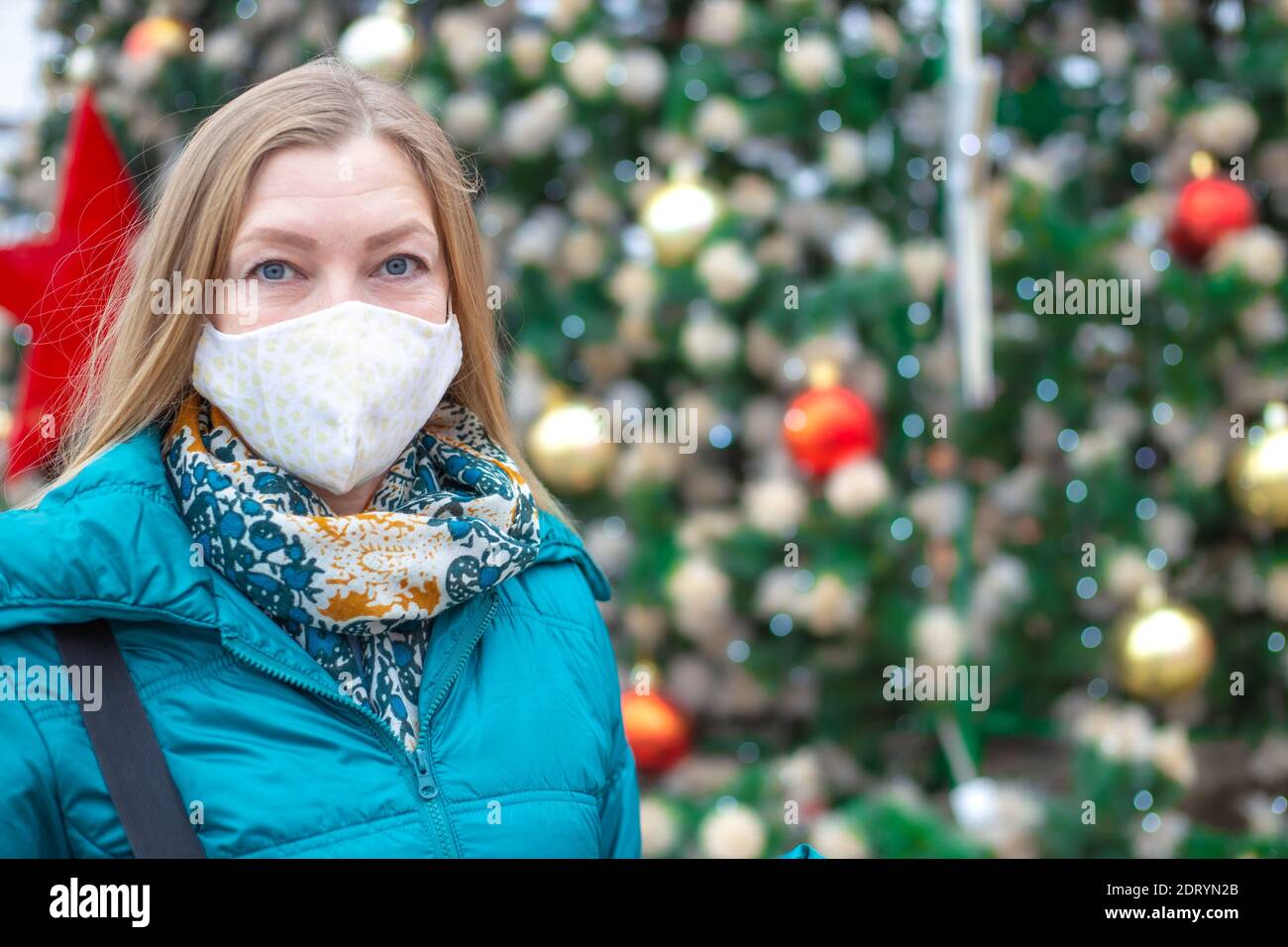 Eine Frau in einer Schutzmaske in der Nähe eines festlich geschmückten Weihnachtsbaums auf der Straße. Weihnachten und Neujahr während der Coronavirus-Pandemie. Stockfoto