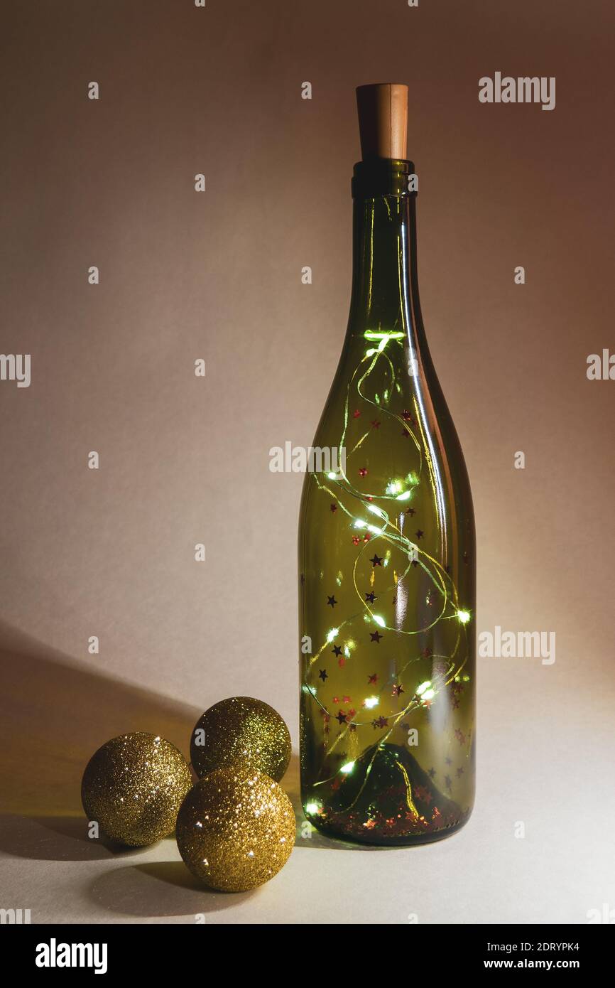 Girlande in Glasflasche für Wein und drei goldglänzende weihnachtskugeln. Selektiver Fokus, vertikale Ansicht Stockfoto