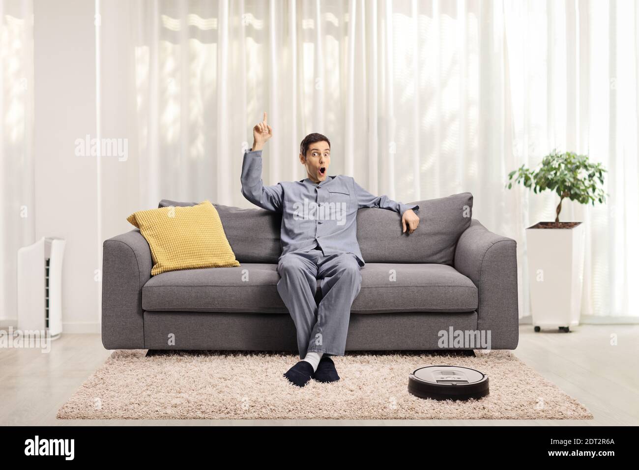 Junge aufgeregt Mann in Pyjamas auf einem Sofa sitzen Nach oben zeigen und ein Roboter-Staubsauger auf dem Boden Stockfoto