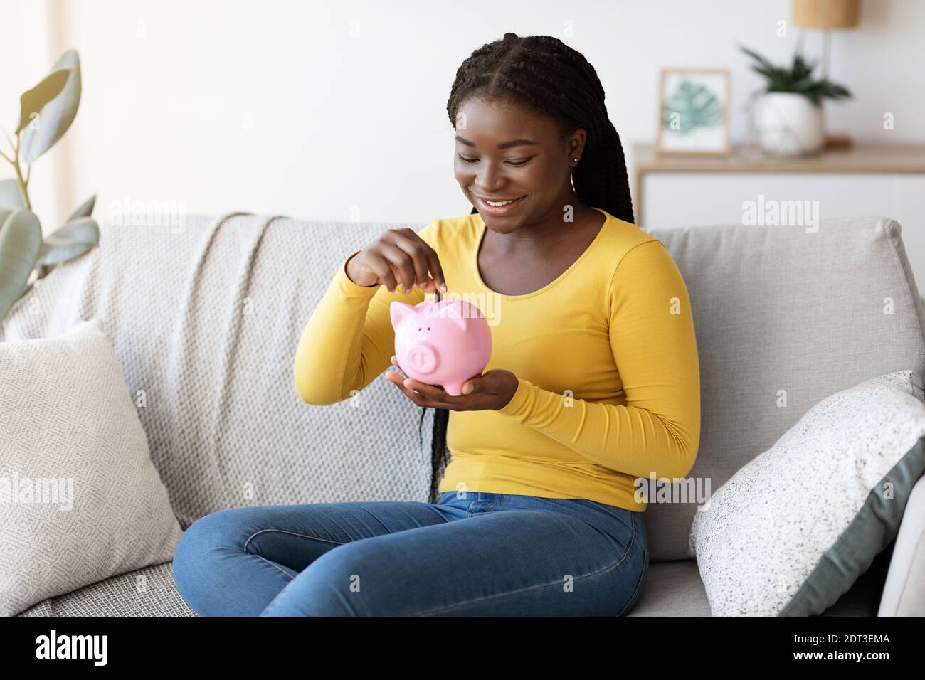 Schwarze Frau, die Münze in Sparschwein legt und zu Hause auf der Couch sitzt Stockfoto