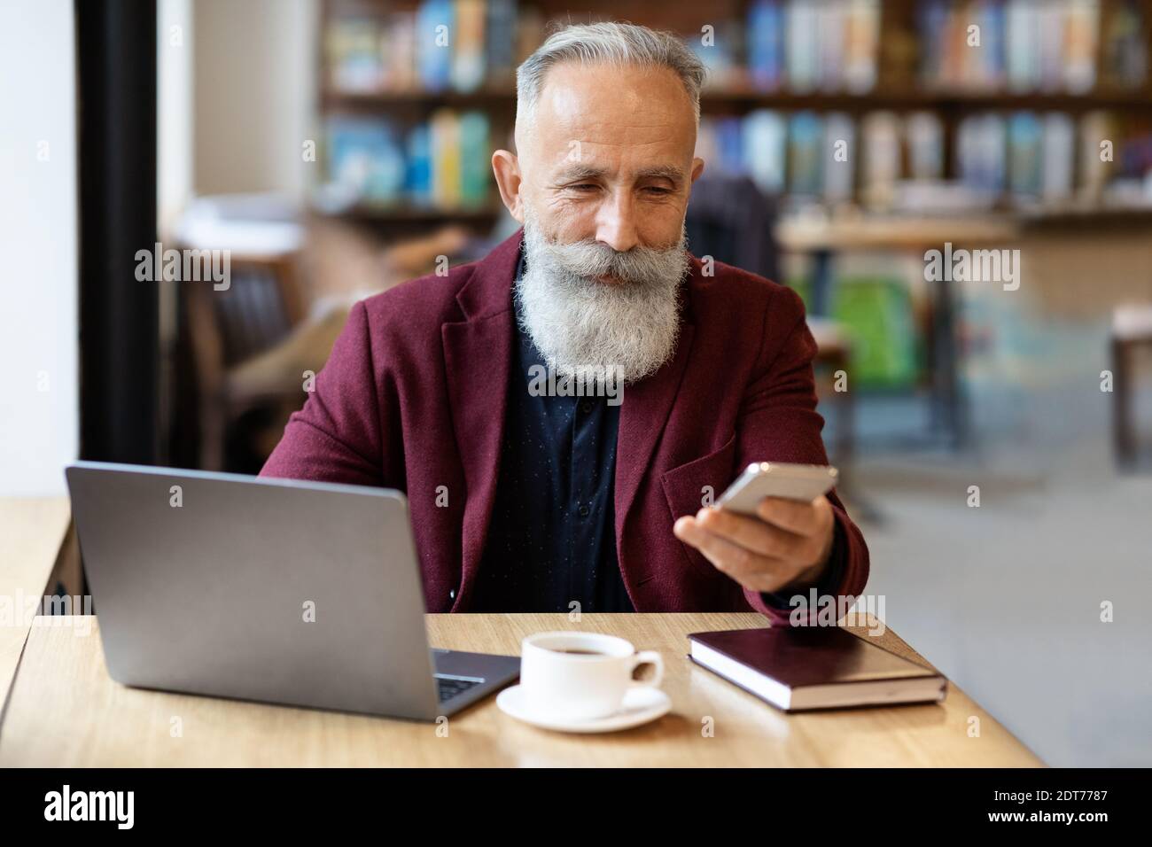 Wohlhabender alter Unternehmer mit Smartphone vor dem Laptop sitzen Stockfoto