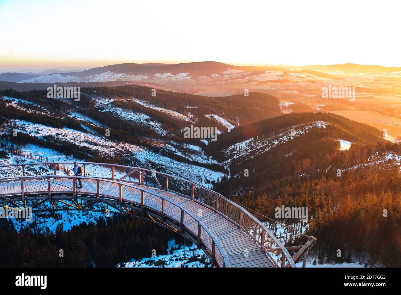 Mann, der auf der hölzernen Brücke des Aussichtsturms Stezka geht v oblacích in Tschechien mit schöner Aussicht auf bedeckte Hügel Mit Schnee bei farbenfrohem Sonnenuntergang Stockfoto