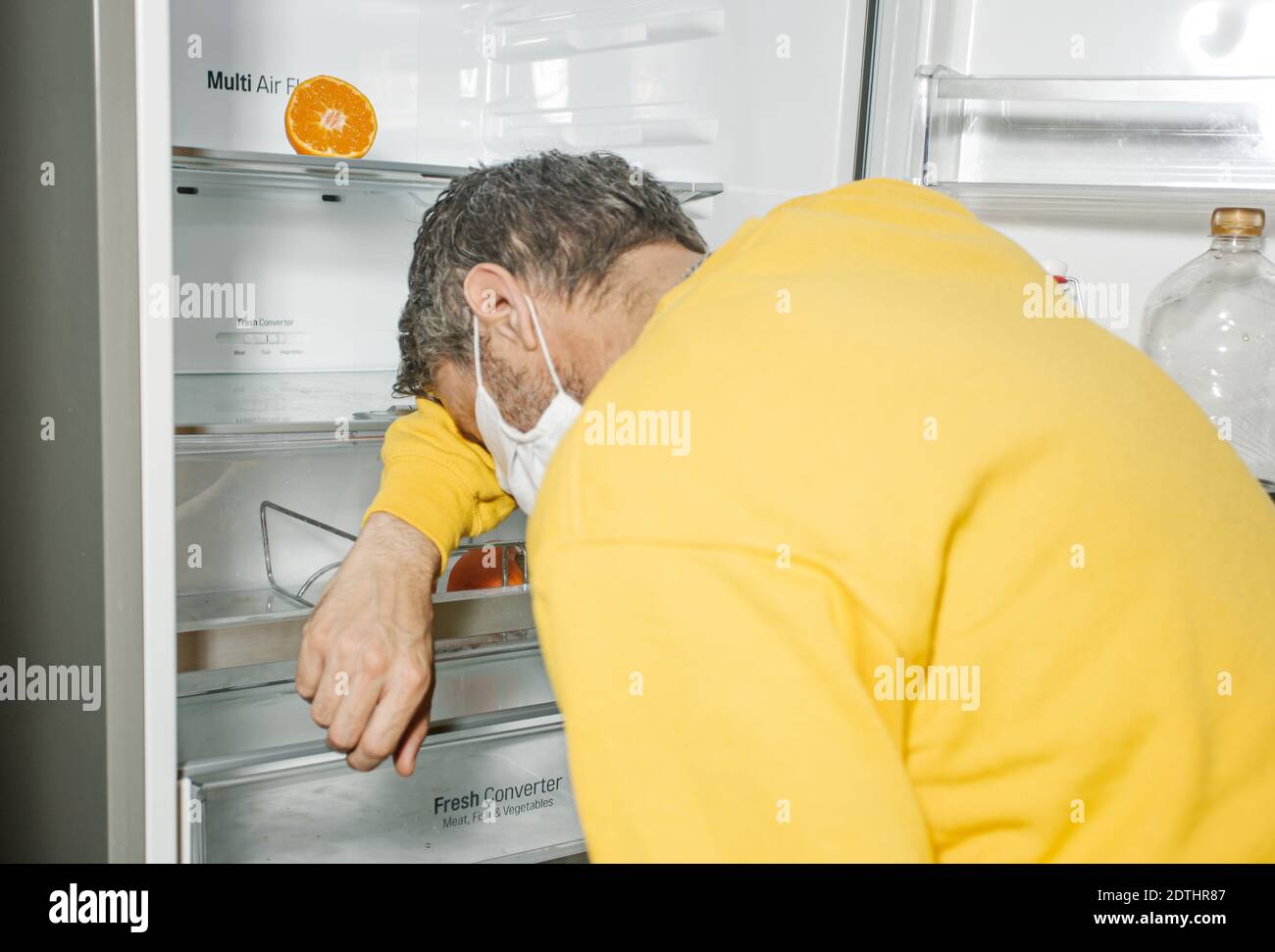 Reifer Mann in Maske, der sich auf die Regale des leeren Kühlschranks lehnte In Erschöpfung während einer Pandemie zu Hause Stockfoto