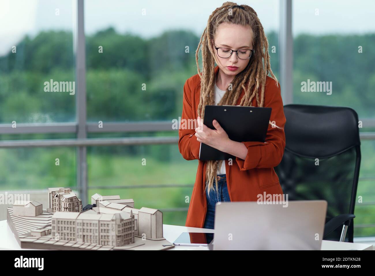 Liebenswert weibliche Ingenieur steht in der Nähe konstruiertes Modell und macht Notizen in die technischen Dokumente. Stockfoto