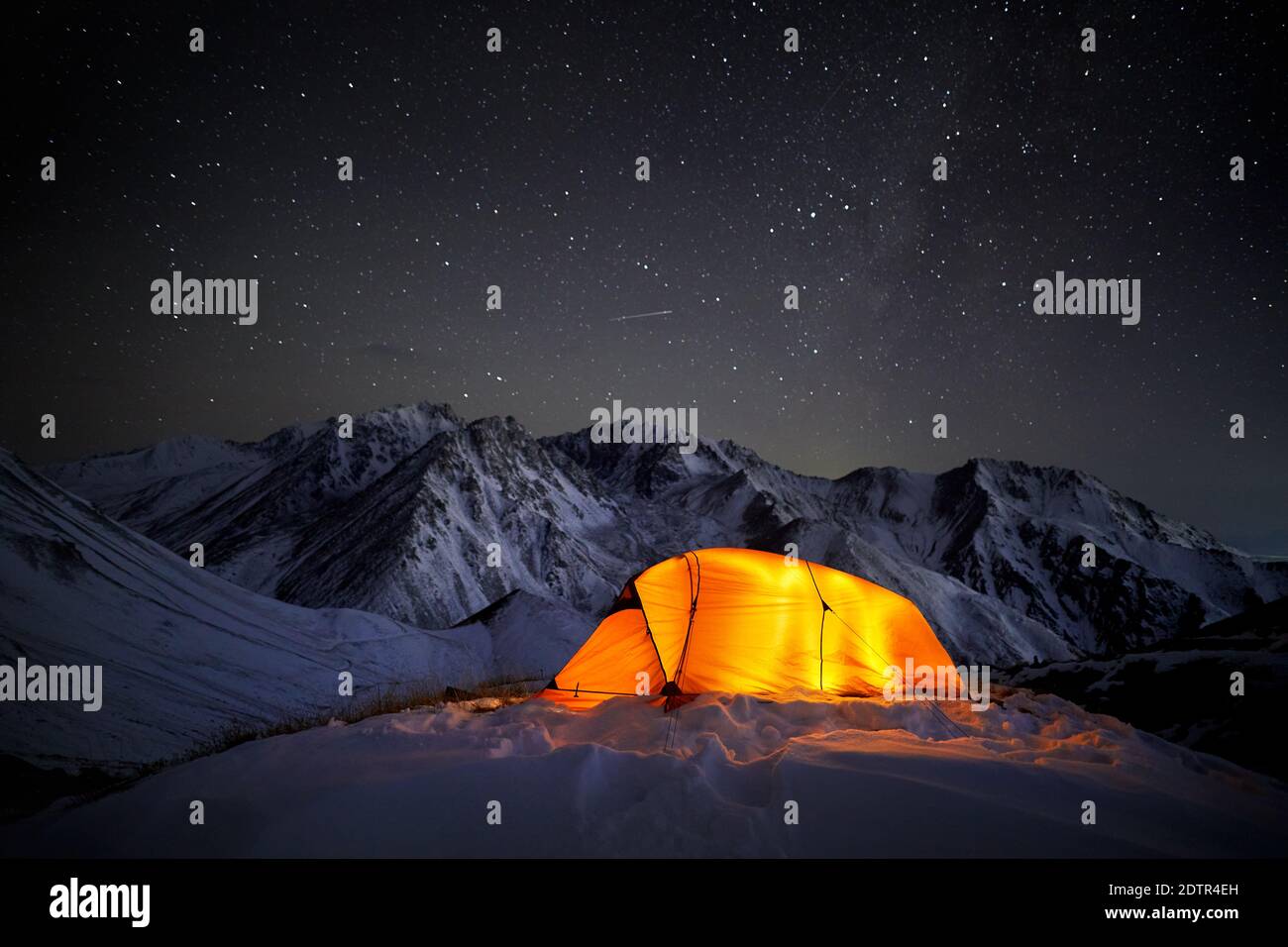 Leuchtendes orangefarbenes Zelt in den Winterbergen bei dunkler Nacht Himmel Hintergrund mit Sternschnuppe Stockfoto