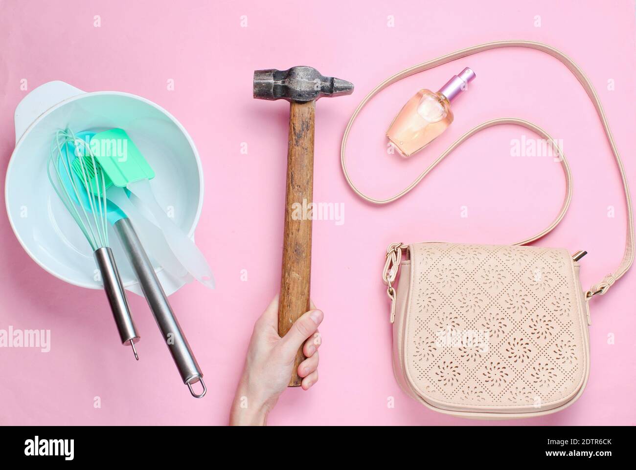 Frauen entscheiden sich für die Arbeit des Mannes. Weibliche Hand hält Hammer auf dem Hintergrund der Schüssel mit Küchengeräten, Taschen und Flasche Parfüm. Rosa Hintergrund, top vie Stockfoto