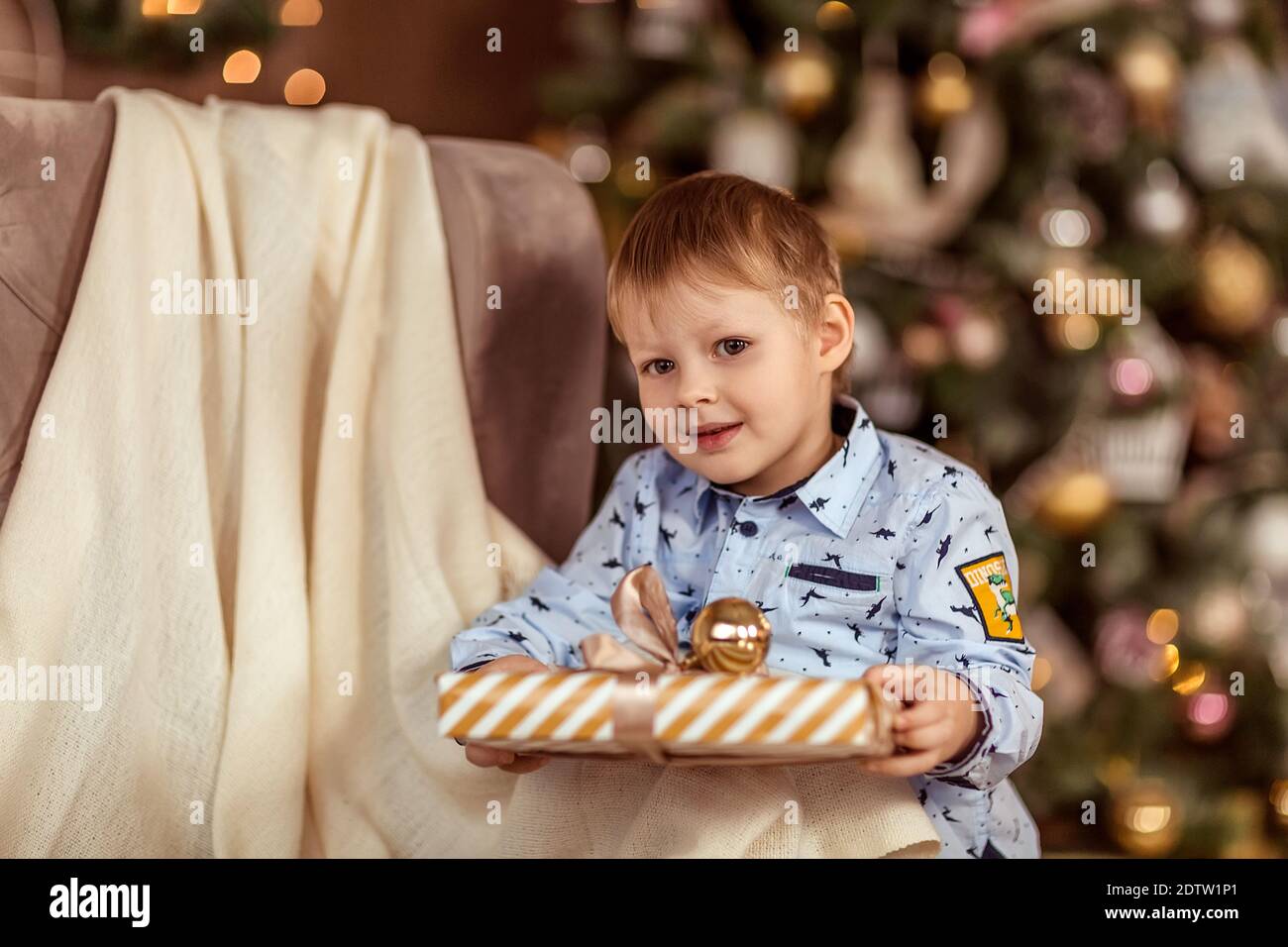 Ein schöner Junge von 4-5 Jahren steht in der Nähe des Sofas und hält Geschenke in seinen Händen. Das Konzept der Winterferien. Selektiver Weichfokus, Bokeh (Unschärfe Stockfoto