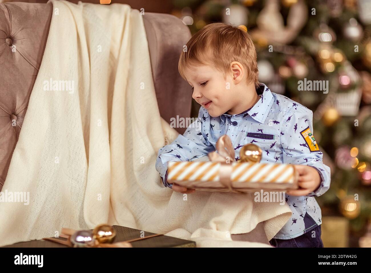 Ein schöner Junge von 4-5 Jahren steht in der Nähe des Sofas und hält Geschenke in seinen Händen. Das Konzept der Winterferien. Selektiver Weichfokus, Bokeh (Unschärfe Stockfoto