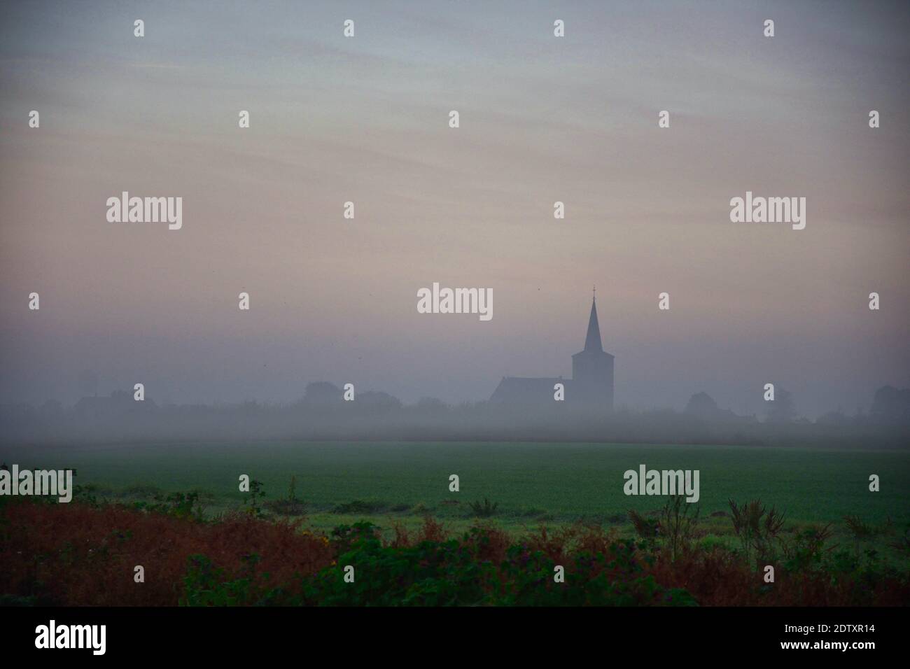 Nebliger Morgen mit Blick auf nasse Felder und vor dem kleinen holländischen Dorf. Morgentau auf dem Land. Alte holländische Kirche im Hintergrund. Stockfoto