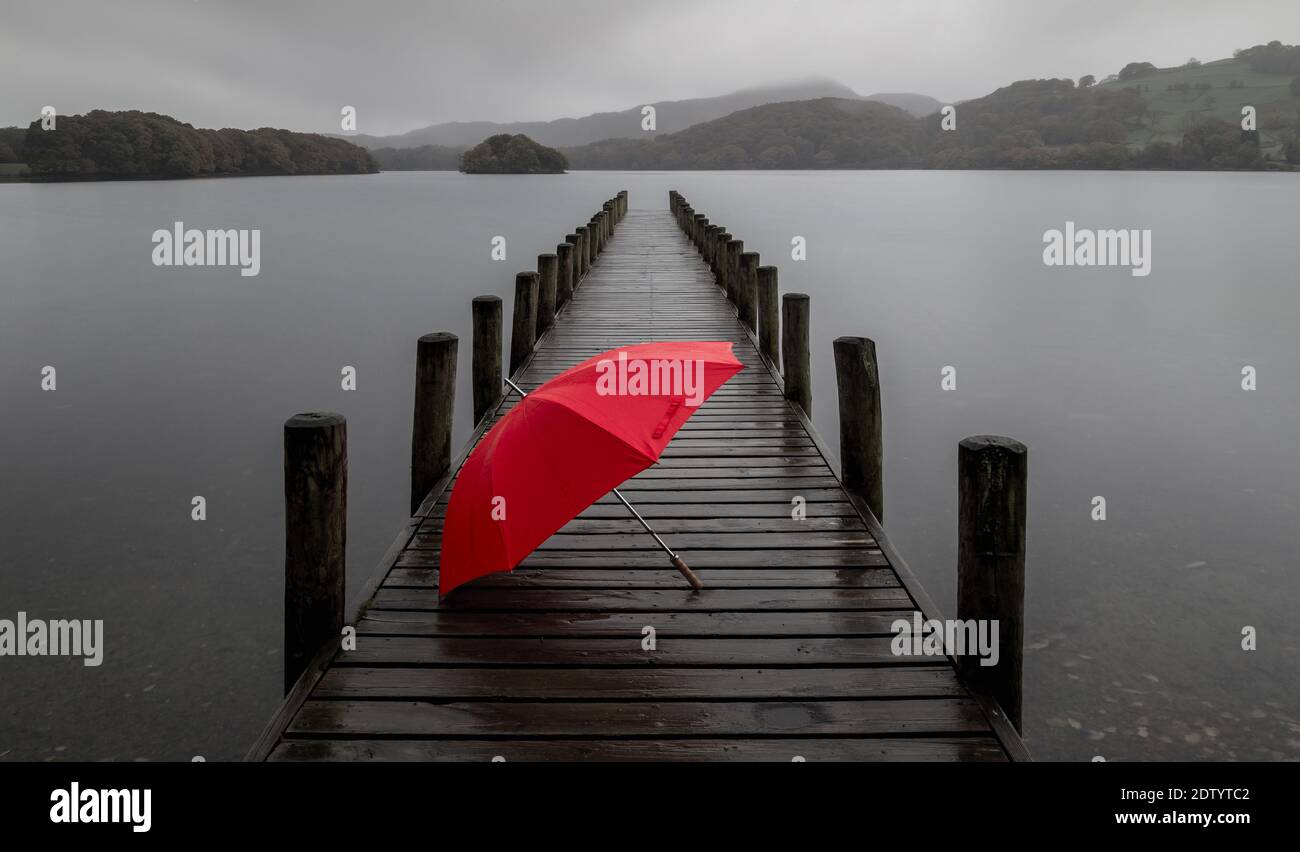 Ein regnerischer grauer Tag am Ufer des Coniston Water, English Lake District, Großbritannien. Der rote Schirm verleiht diesem fast monochromen Foto einen Farbtupfer. Stockfoto