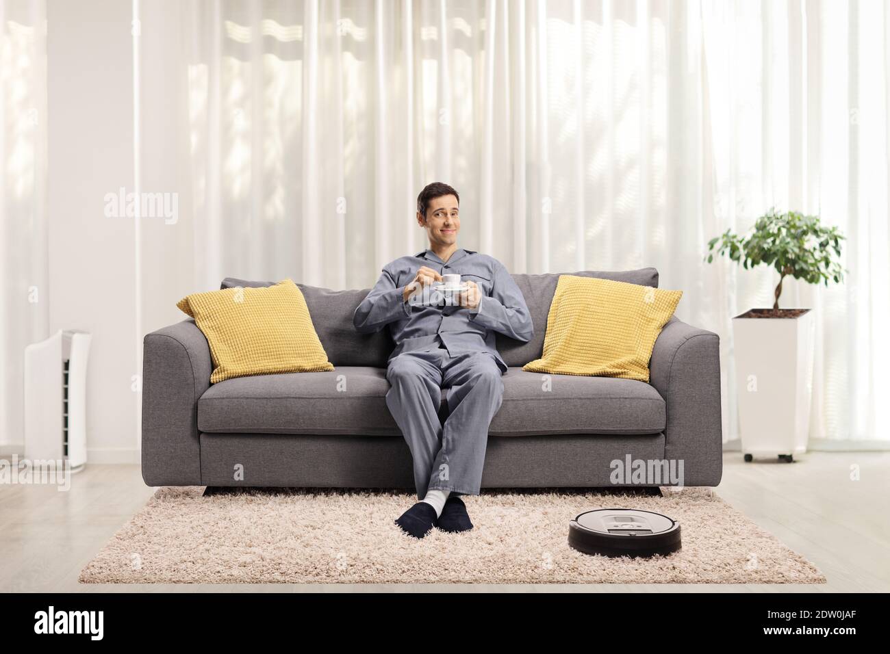 Roboter-Staubsauger auf dem Teppich und ein Mann in Schlafanzug auf dem Sofa mit einer Tasse Espresso Stockfoto