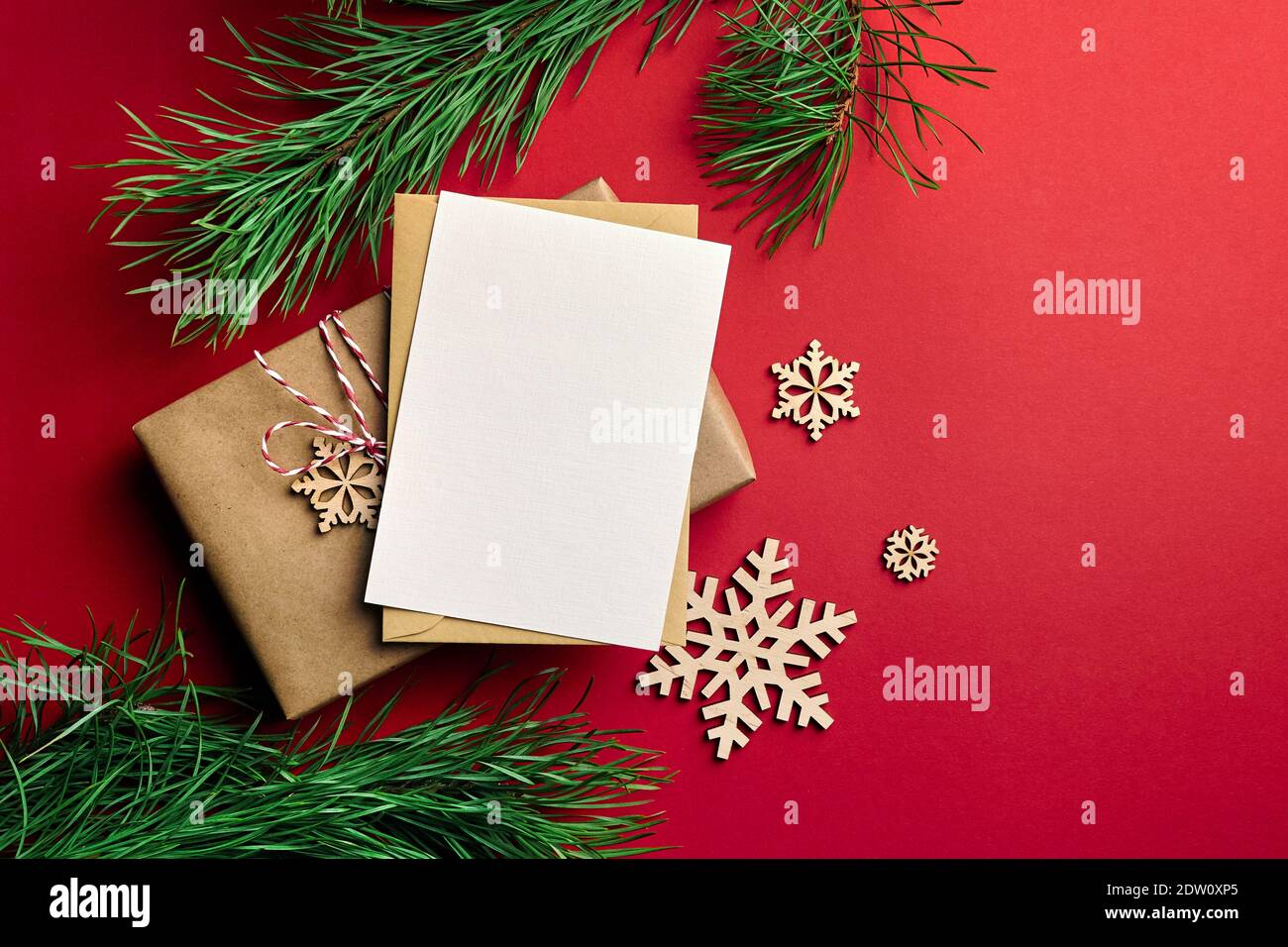Weihnachten Grußkarte mockup. Geschenkbox, hölzerne Schneeflocken Dekorationen und Kiefernäste auf rotem Papier Hintergrund. Draufsicht, Kopierbereich. Stockfoto