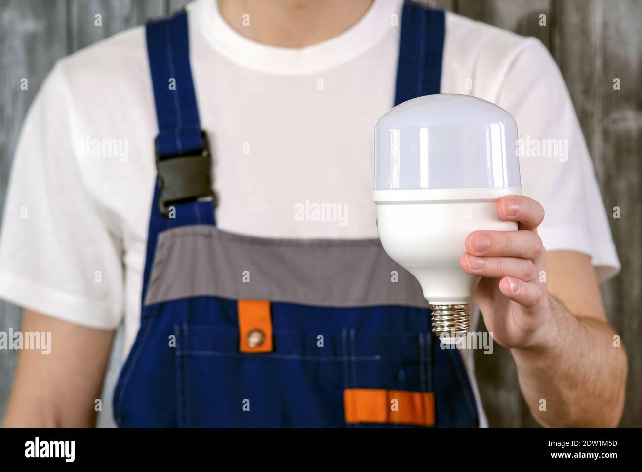 Ein Elektriker in einem blauen Jumpsuit. In der Hand hält er eine leistungsstarke industrielle Energiesparlampe. Konzept der Energieeinsparung Stockfoto