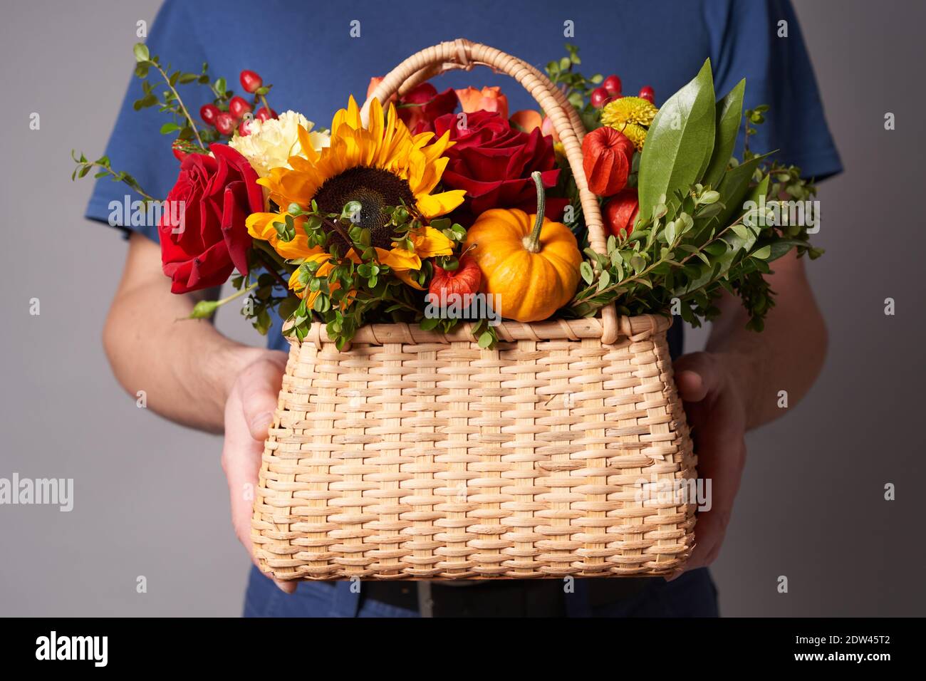 Ein weißer Mann in einem blauen T-Shirt hält einen Blumenkorb mit einem Kopierplatz auf einem grauen Hintergrund, Blumenlieferung oder ein Geschenk für den Urlaub Stockfoto