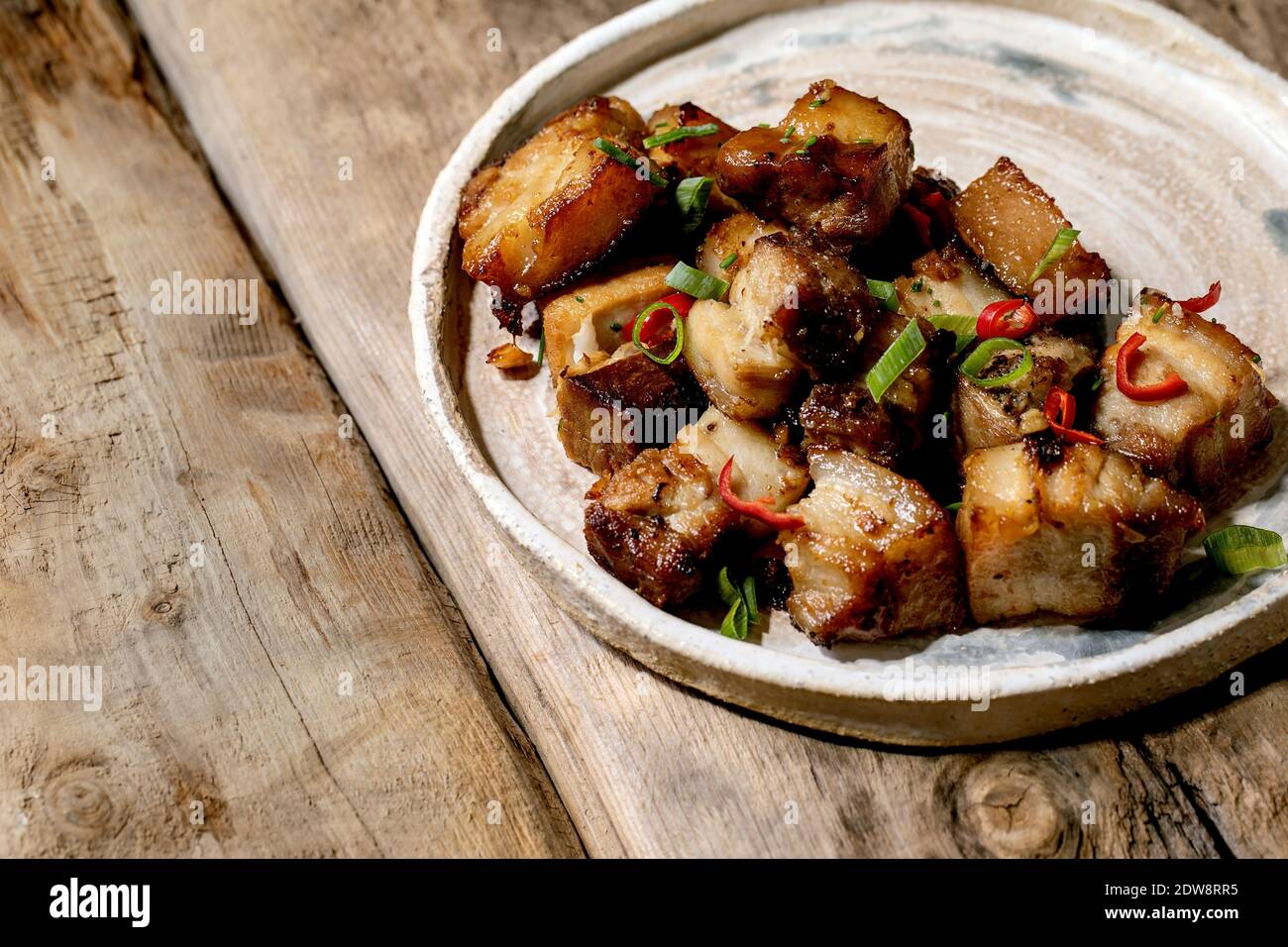 Gegrillter frittierter Schweinebauch mit Chili-Pfeffer und Frühlingszwiebeln auf Keramikplatte mit Essstäbchen auf altem Holzhintergrund. Flach liegend, Kopierplatz Stockfoto