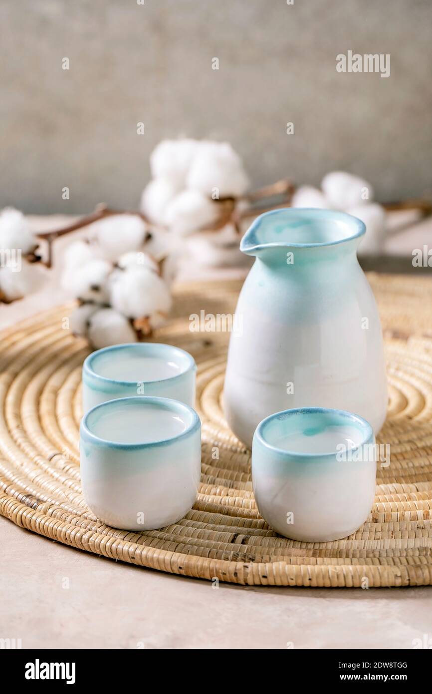 Sake Keramik Set für traditionelle japanische Alkohol trinken Reiswein Sake, Krug und drei Tassen, stehend auf Strohserviette mit Baumwollblumen über beig Stockfoto