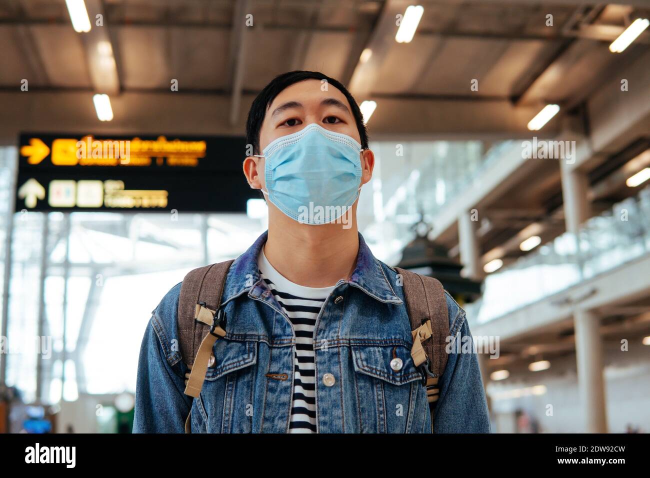 Asiatischer männlicher Reisender, der eine Gesichtsmaske trägt, die auf das Einsteigen in ein Flugzeug wartet. Junge Touristen warten auf Flughafen mit Abflug Bord im Hintergrund während Covid-19 Virus Pandemie Stockfoto